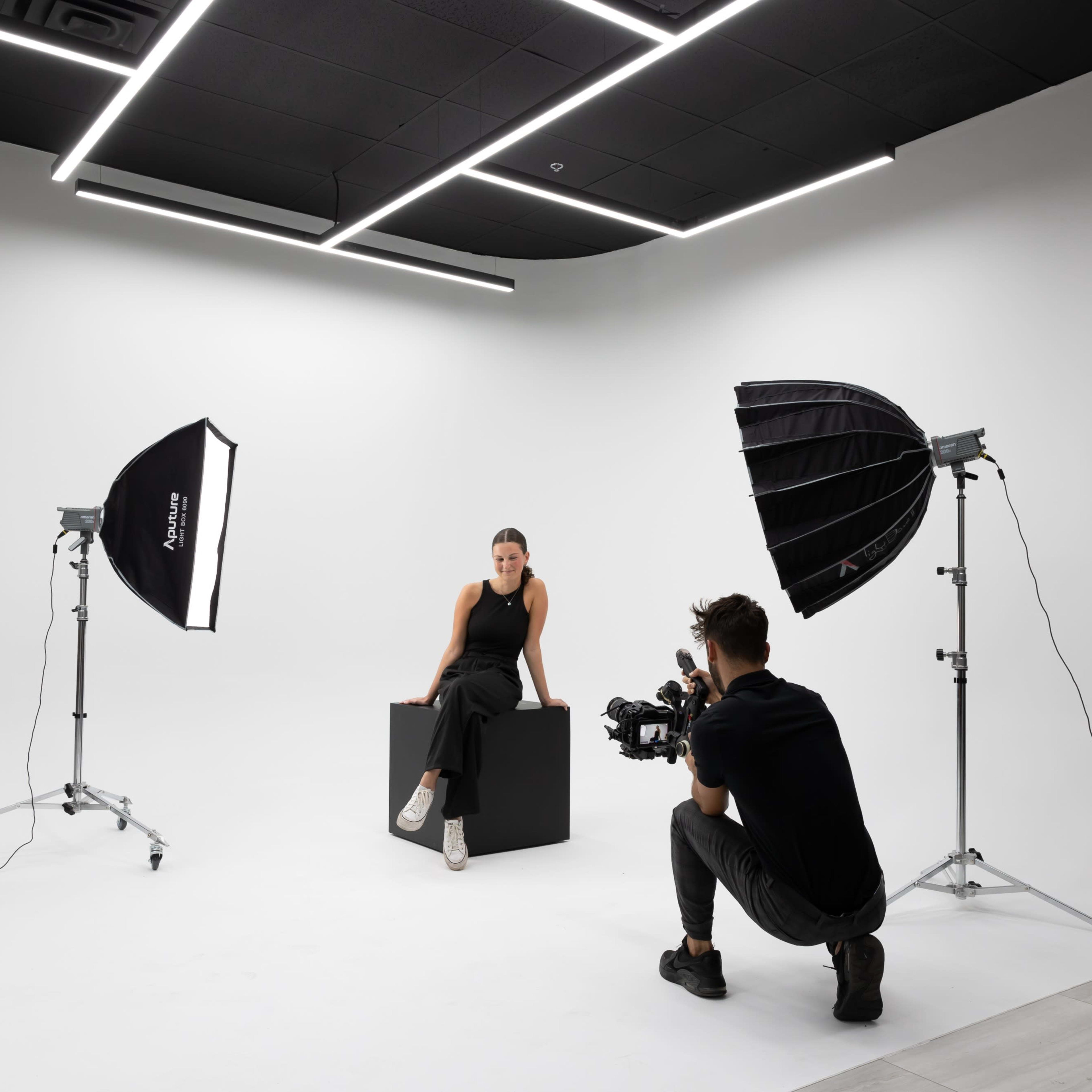 A photographer captures a model sitting on a black cube in a studio with a white backdrop and two softbox lights.