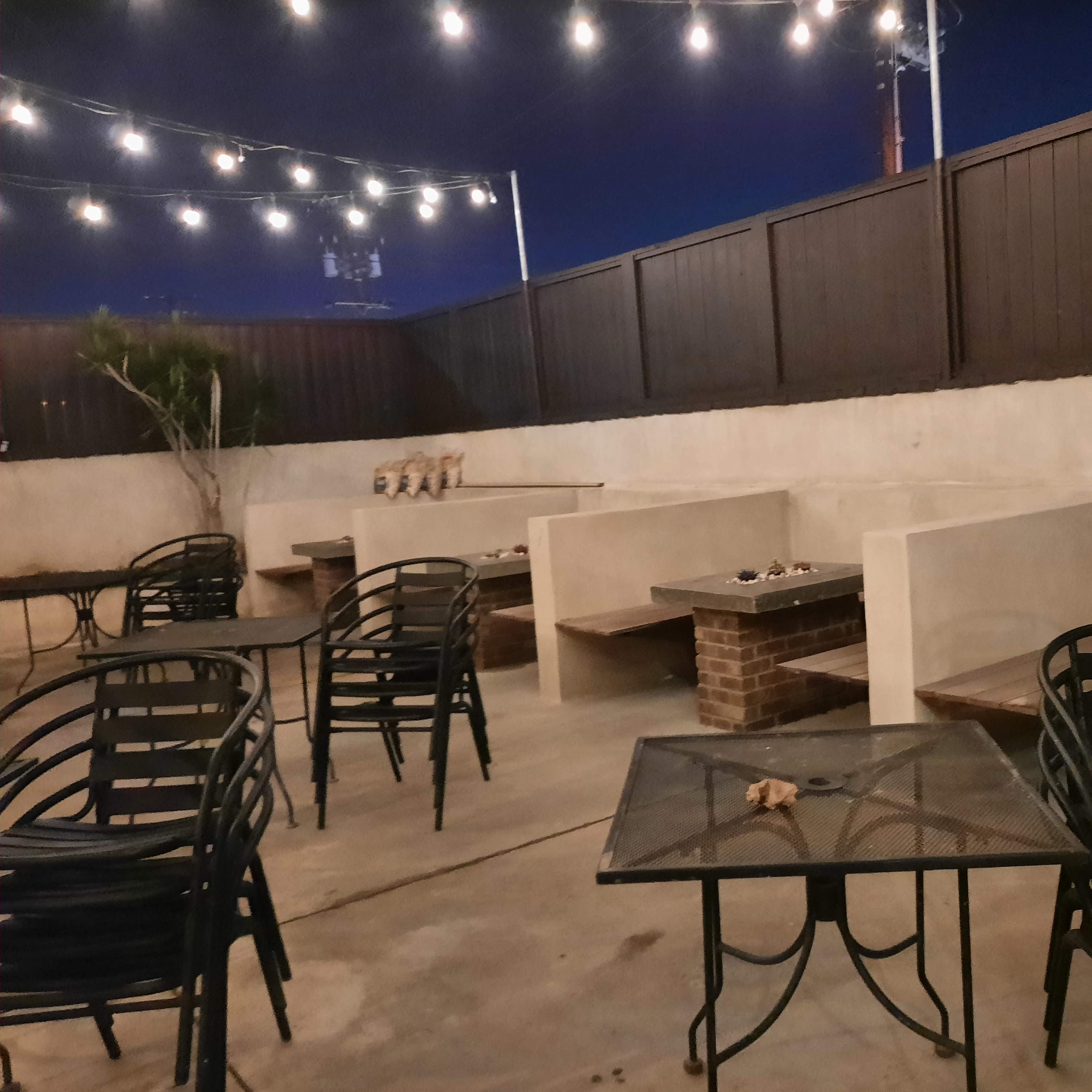A dimly lit outdoor seating area features a row of benches and tables surrounded by string lights in the evening.