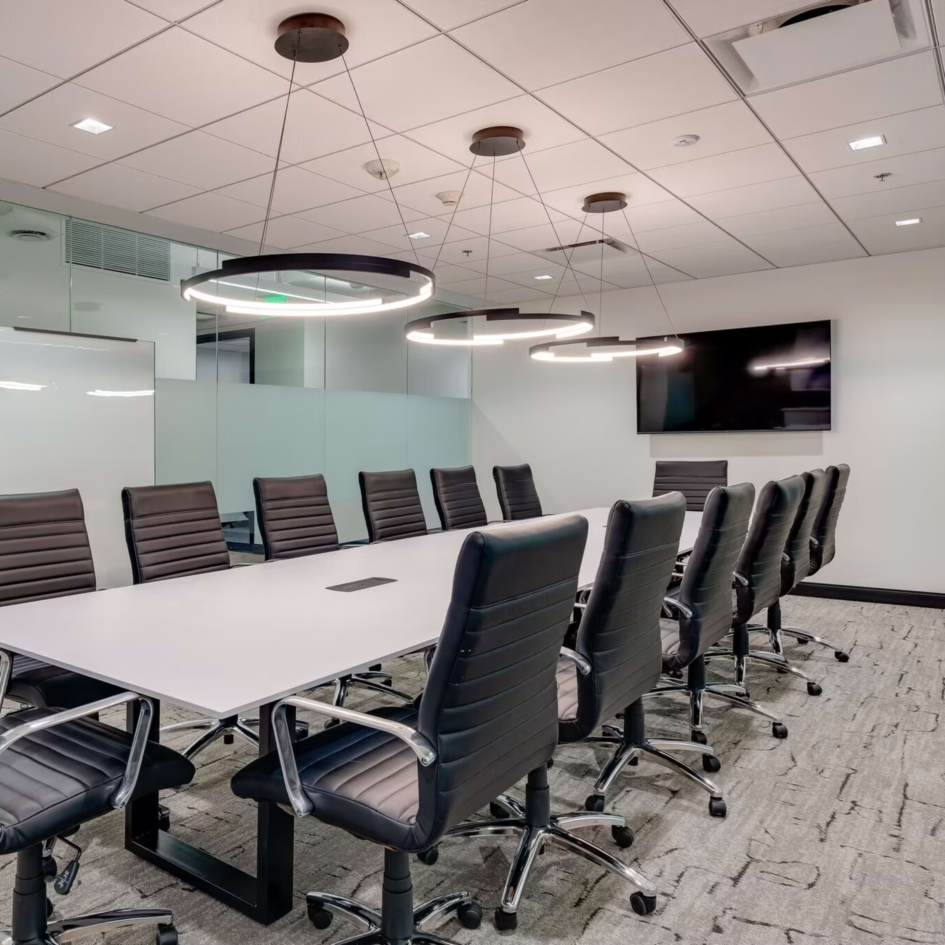A modern conference room features a large table surrounded by black ergonomic chairs, with glass walls and circular light fixtures.