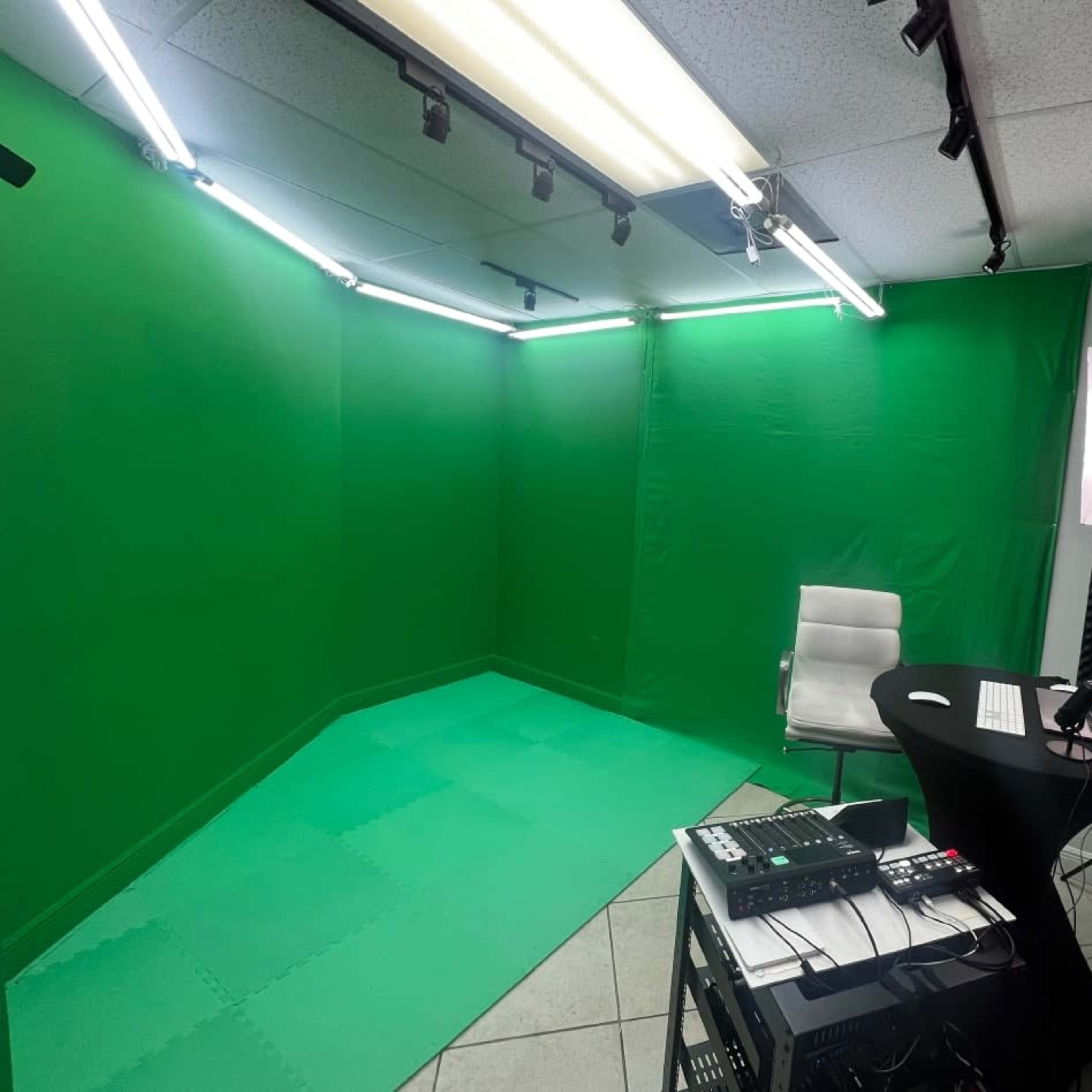 A green screen studio set up with a white chair, a black desk with a computer, and various recording equipment.