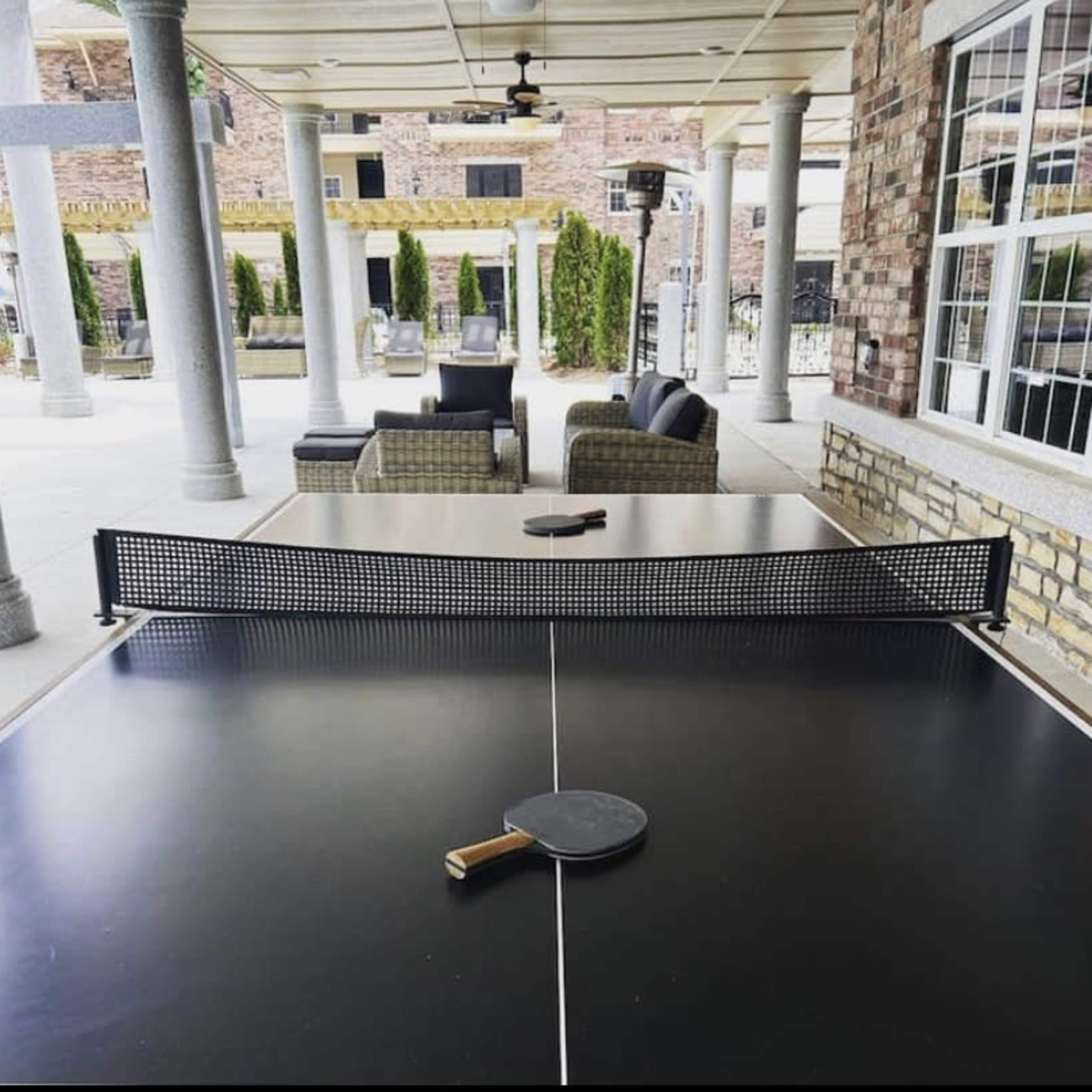 A ping pong table sits under a covered patio, surrounded by seating and decorative columns.
