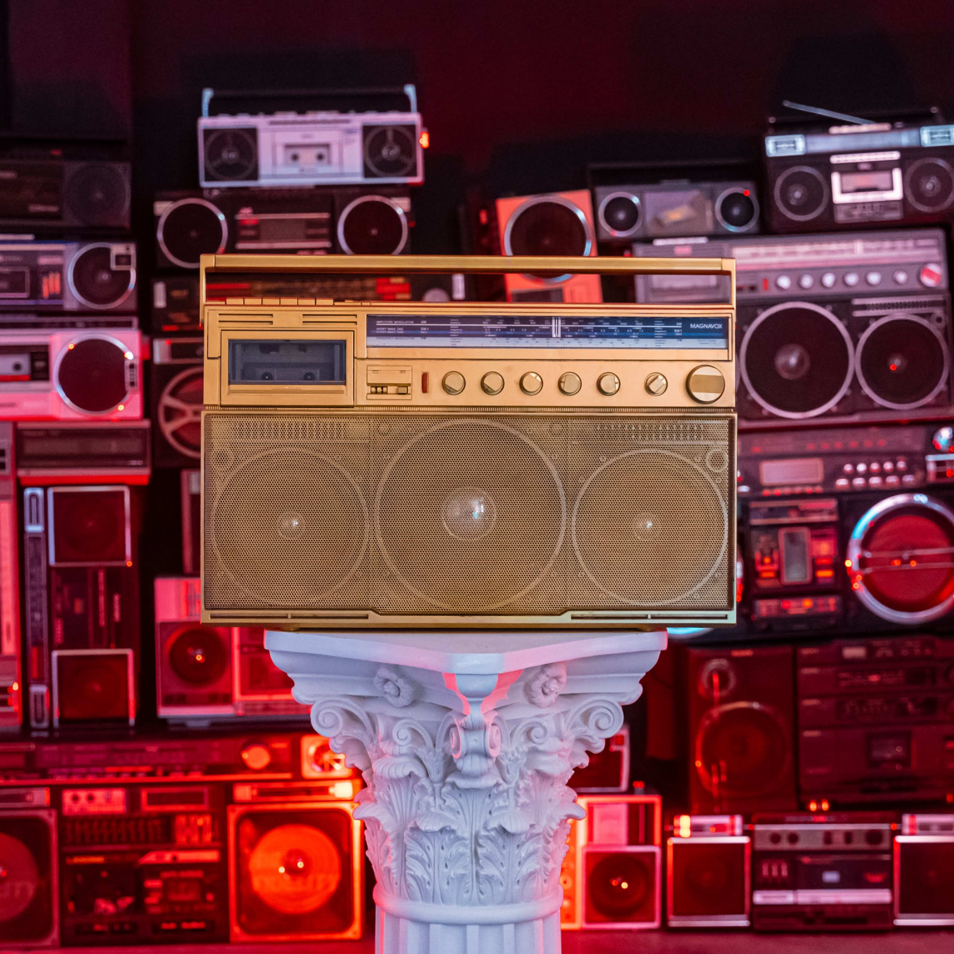 A vintage boombox is displayed on a prominent pedestal in front of a wall stacked with various cassette players and radios.