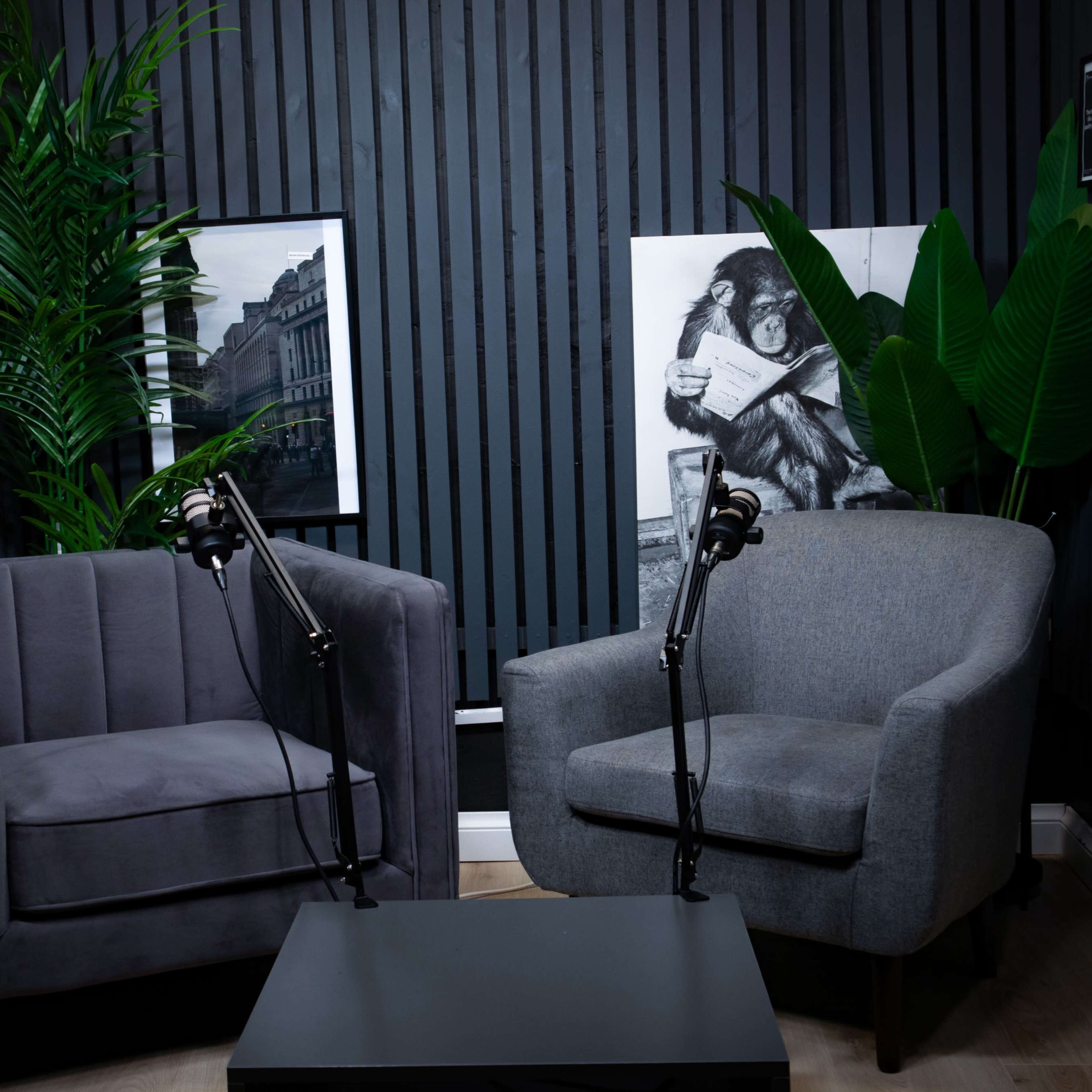 A podcast recording setup featuring two armchairs with microphones on stands, surrounded by framed artwork and greenery on a striped wall.