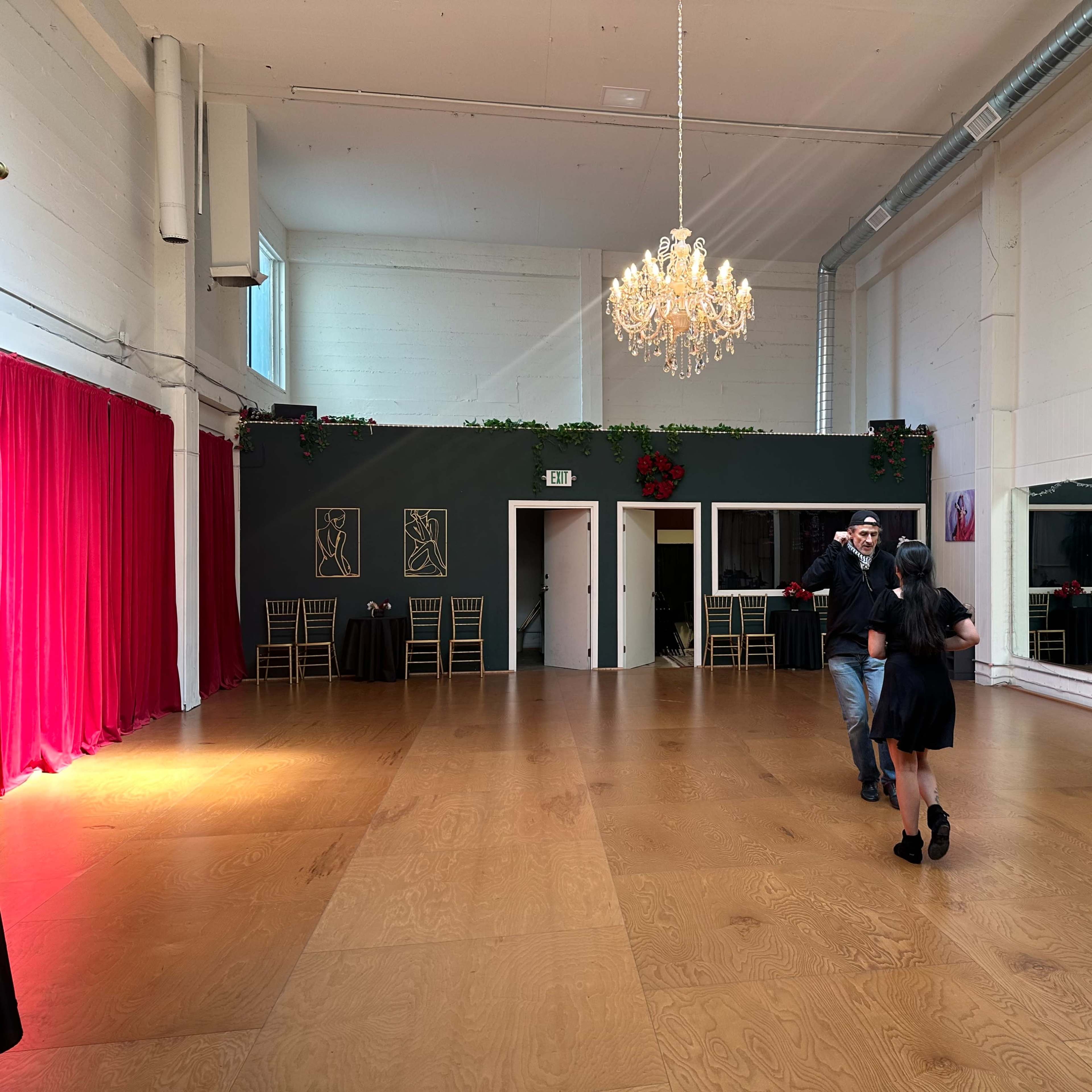 A spacious dance studio features a wooden floor, large mirrors, and a chandelier, with two people dancing in the center.