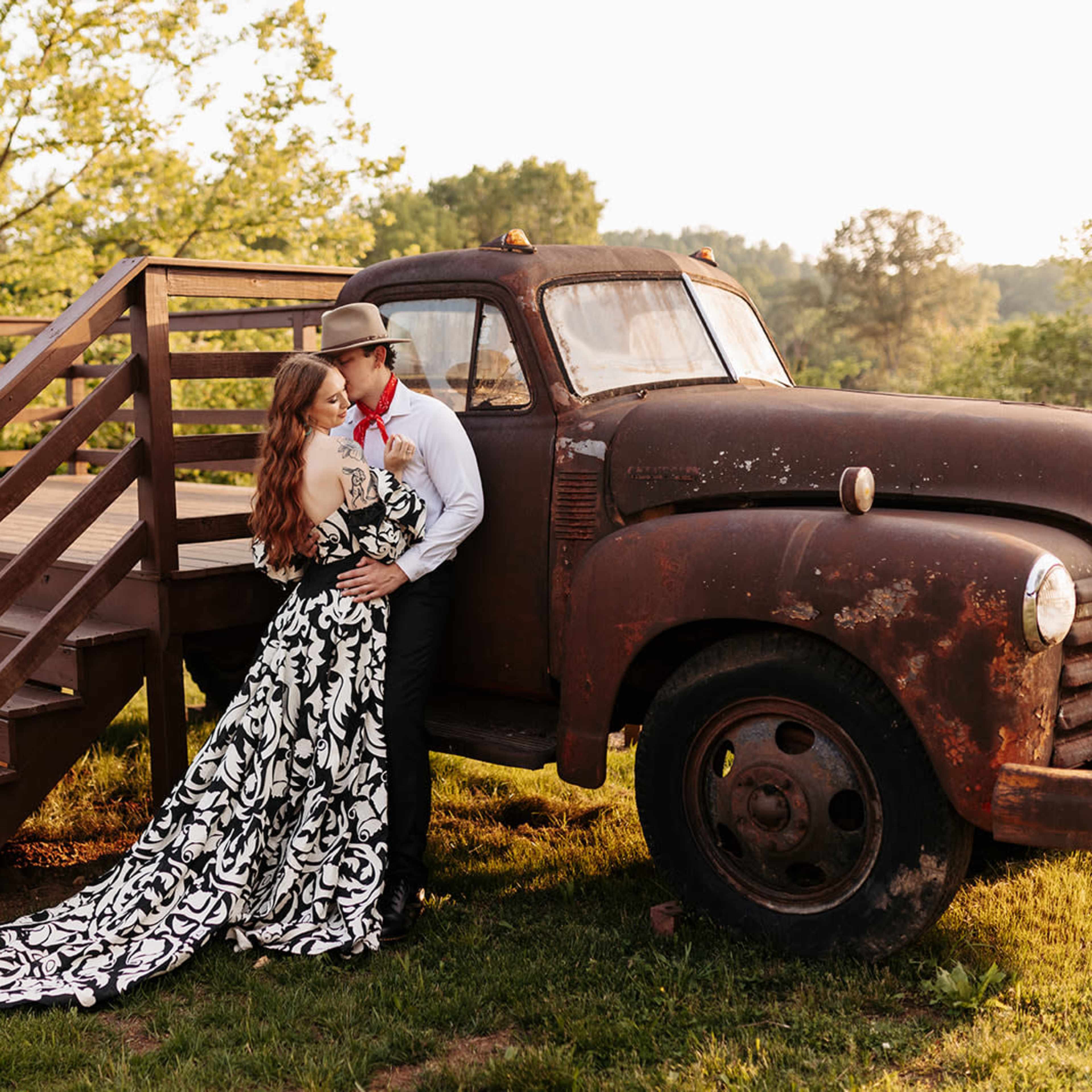 A couple poses near an old, rusty pickup truck next to a wooden staircase in a lush green outdoor setting.