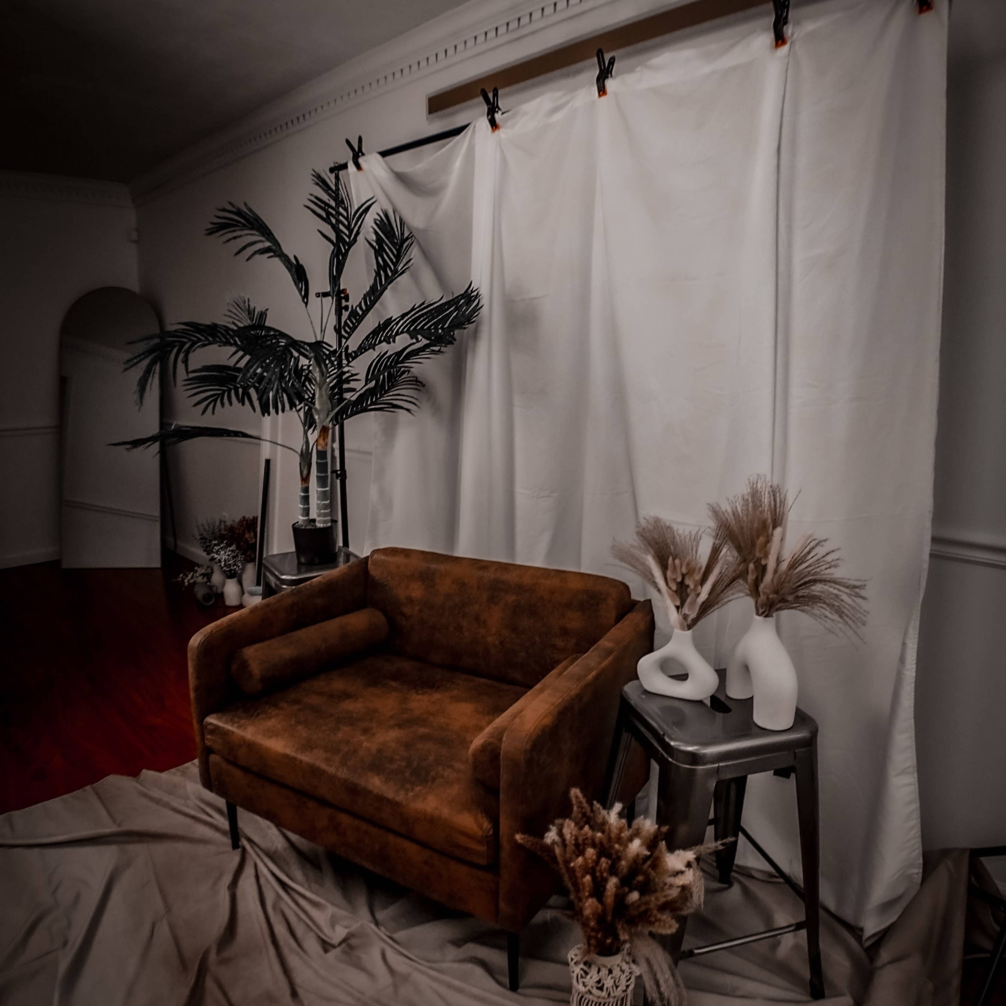 A brown couch is placed in front of a white backdrop, accompanied by decorative vases and dried plants on a small table.