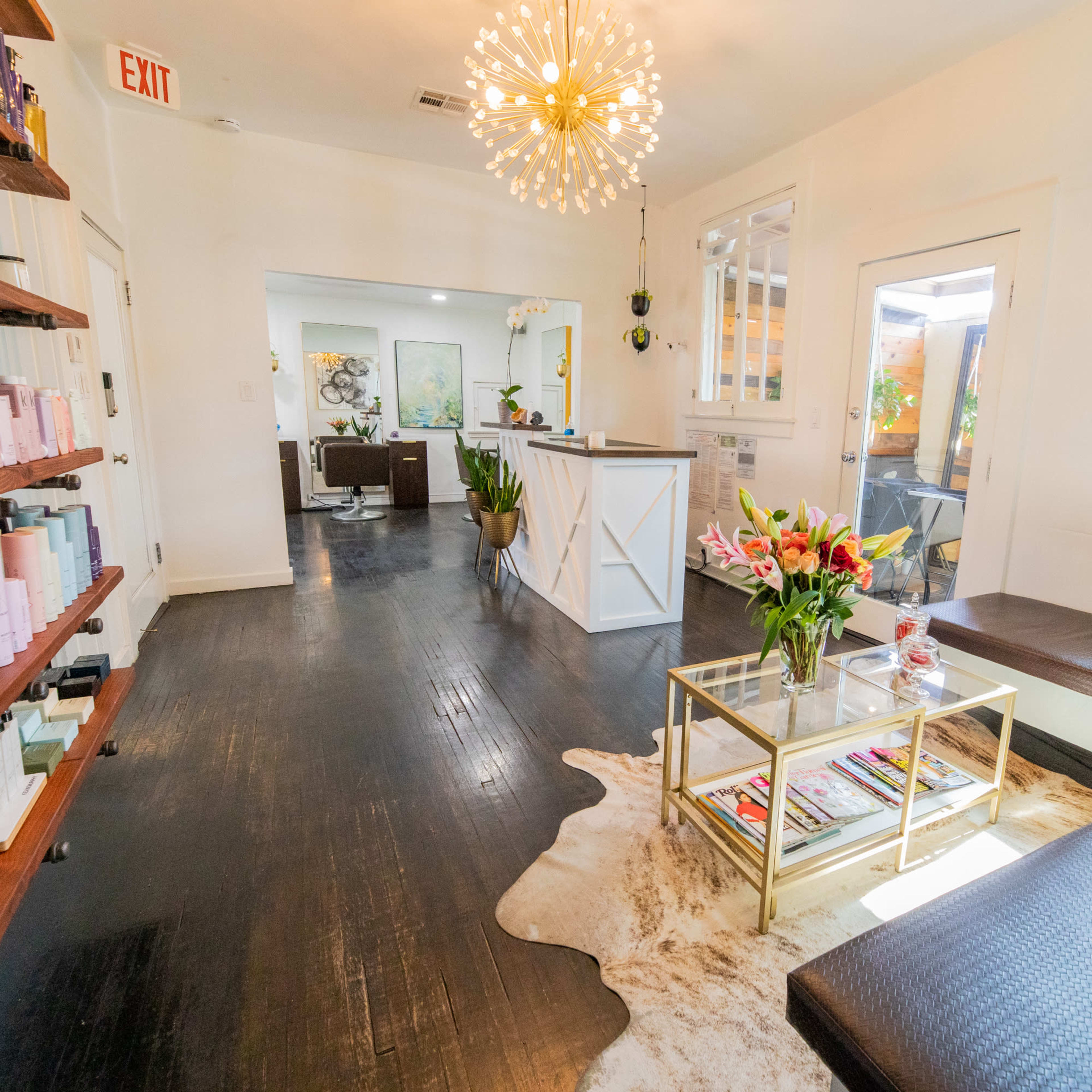 A bright, modern salon interior with a seating area featuring a glass coffee table, a large decorative light fixture, and shelves stocked with beauty products.
