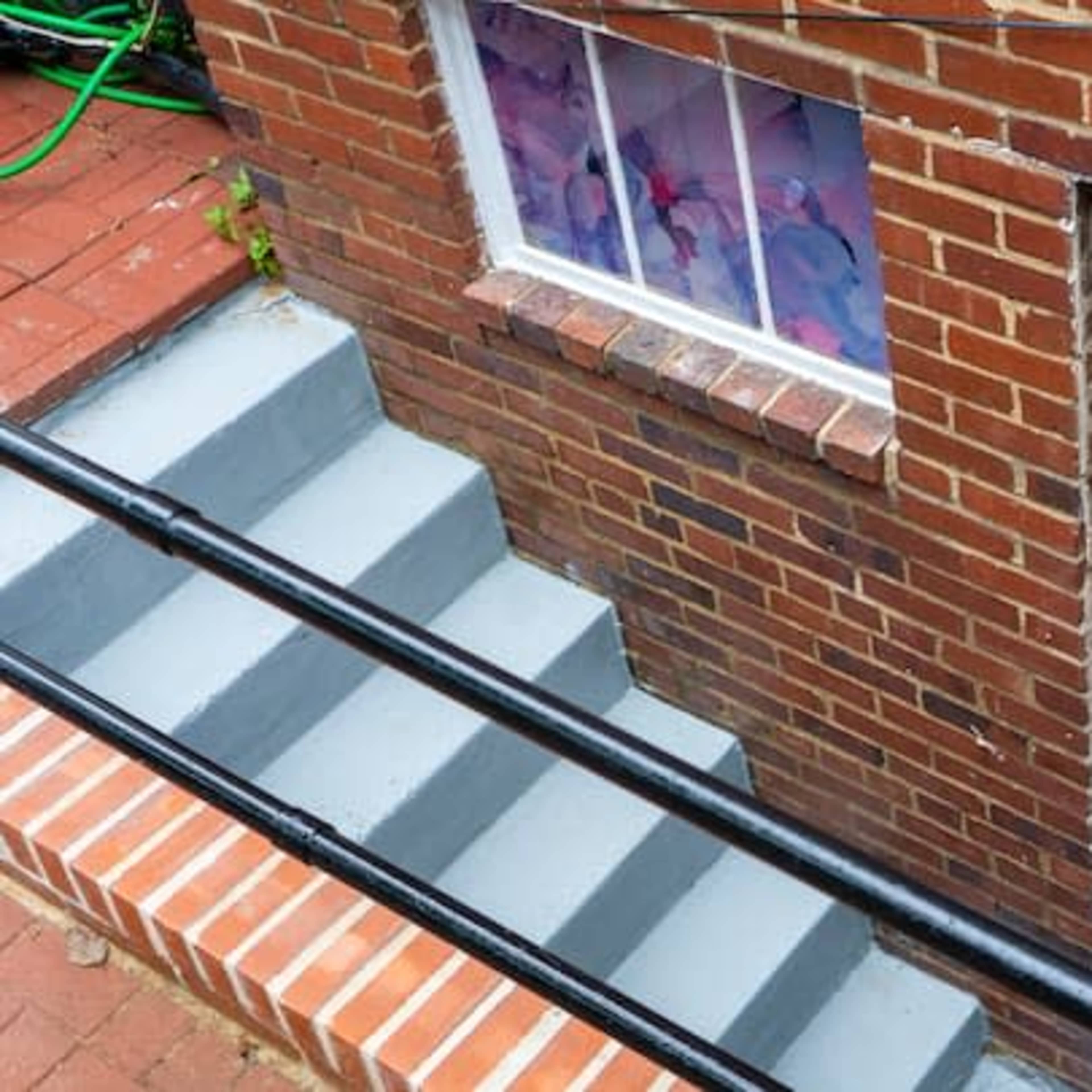 A brick exterior of a house with a set of gray stairs leading down to a white door, featuring a stained glass window, and a small window above it.