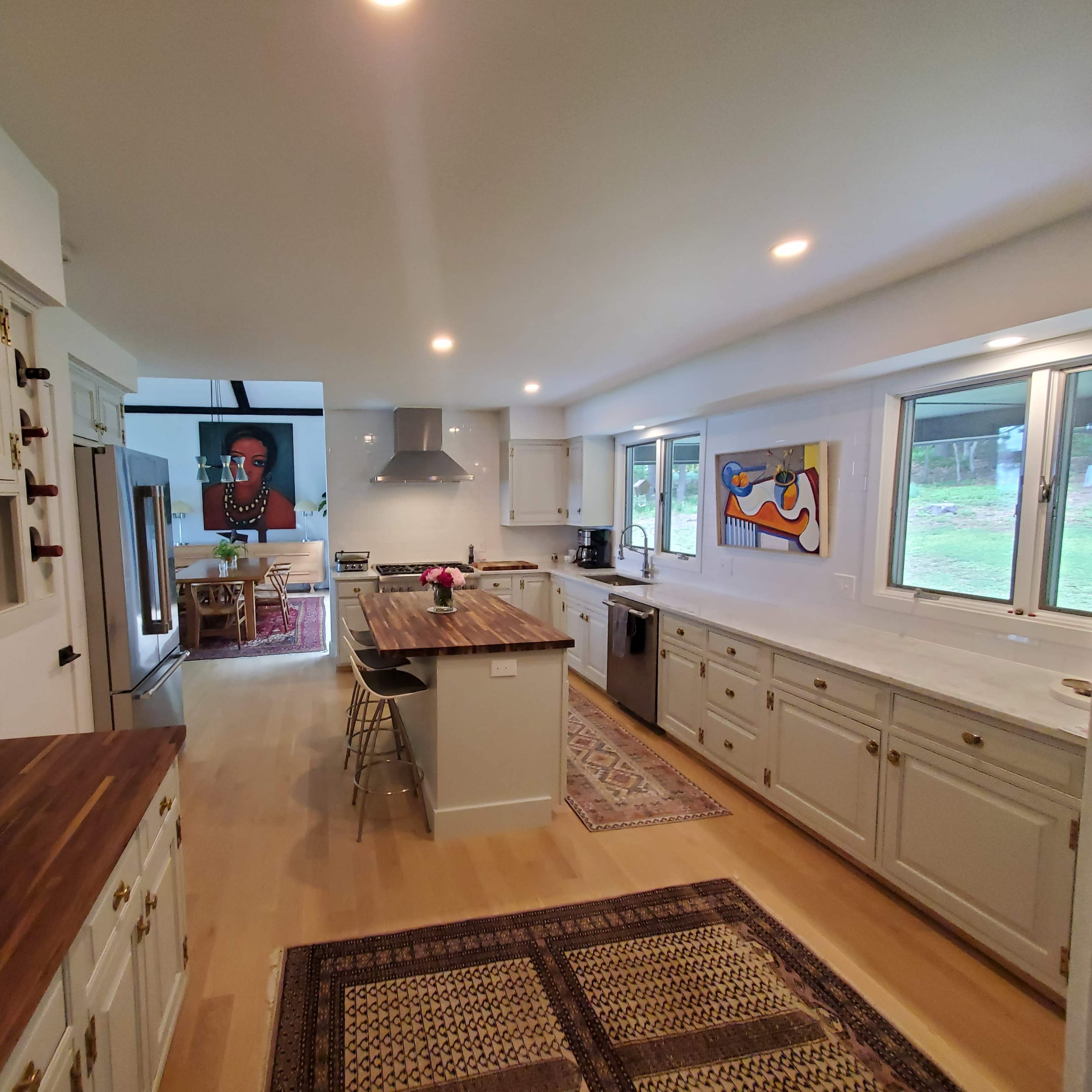 A bright kitchen features white cabinetry, a central island with a countertop, and large windows overlooking a lush outdoor area.