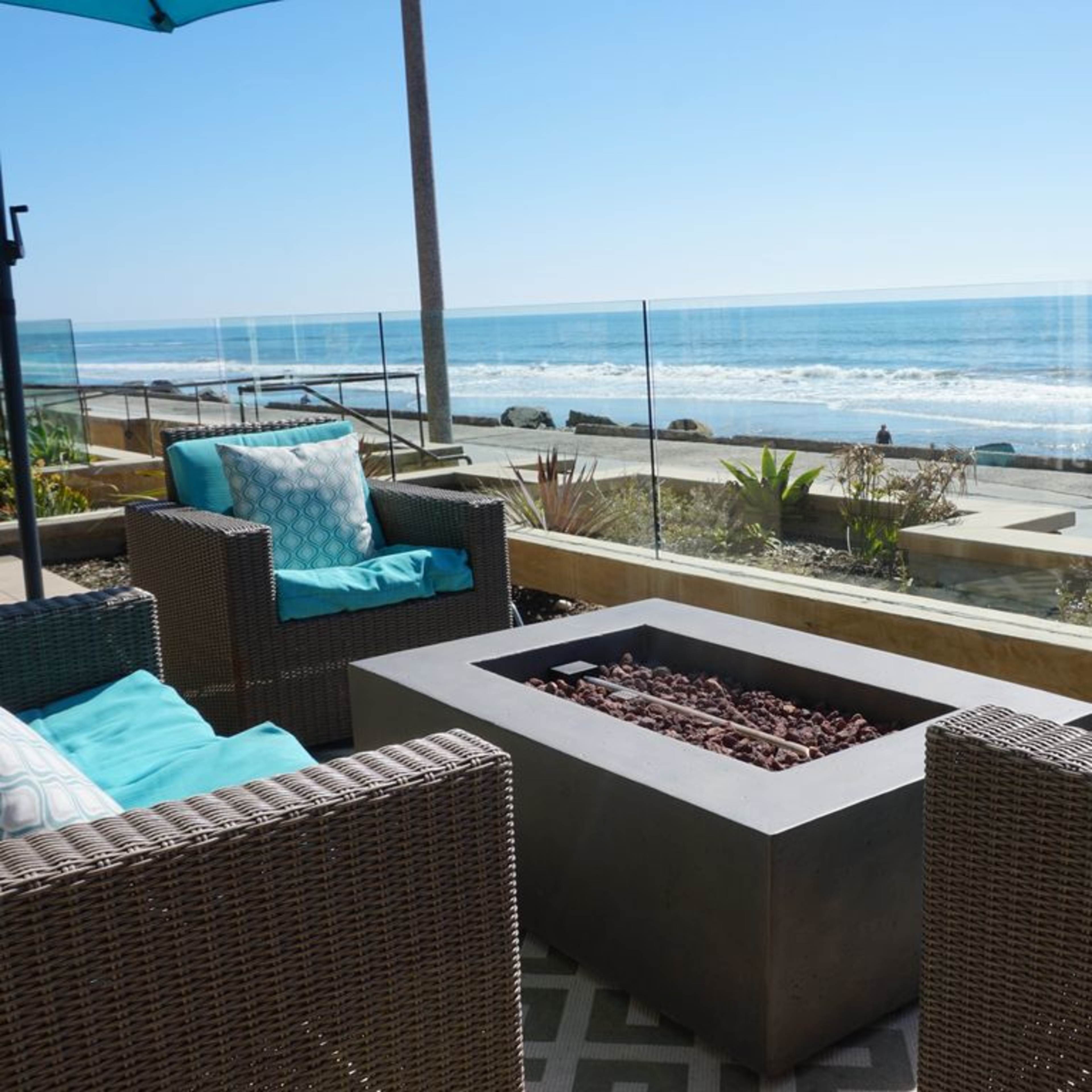 A modern outdoor seating area features two wicker chairs with blue cushions, surrounding a rectangular fire pit, overlooking the ocean.