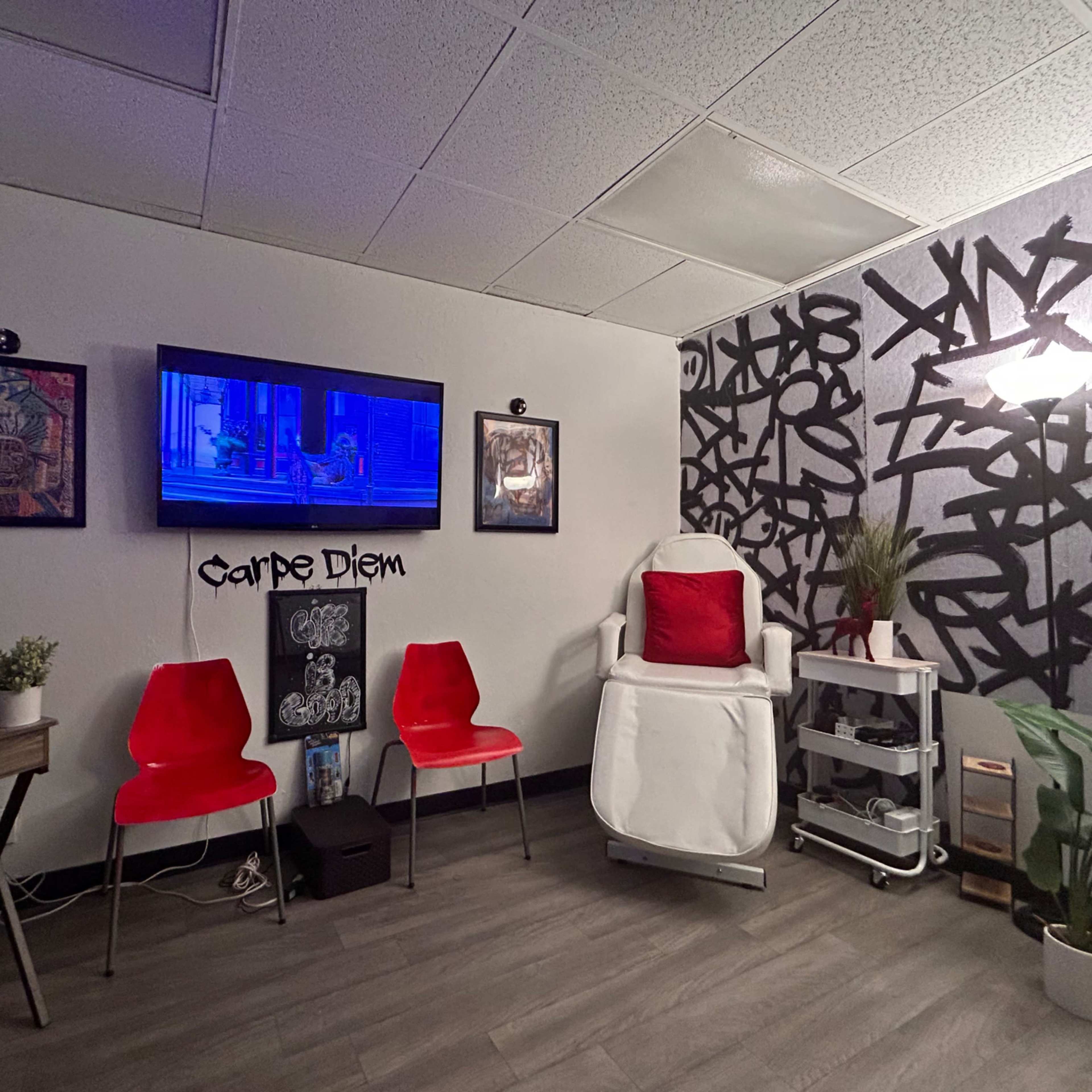 A small, modern room with a reclined chair, a television on the wall, and two red chairs in front of a decorated wall featuring black patterns.
