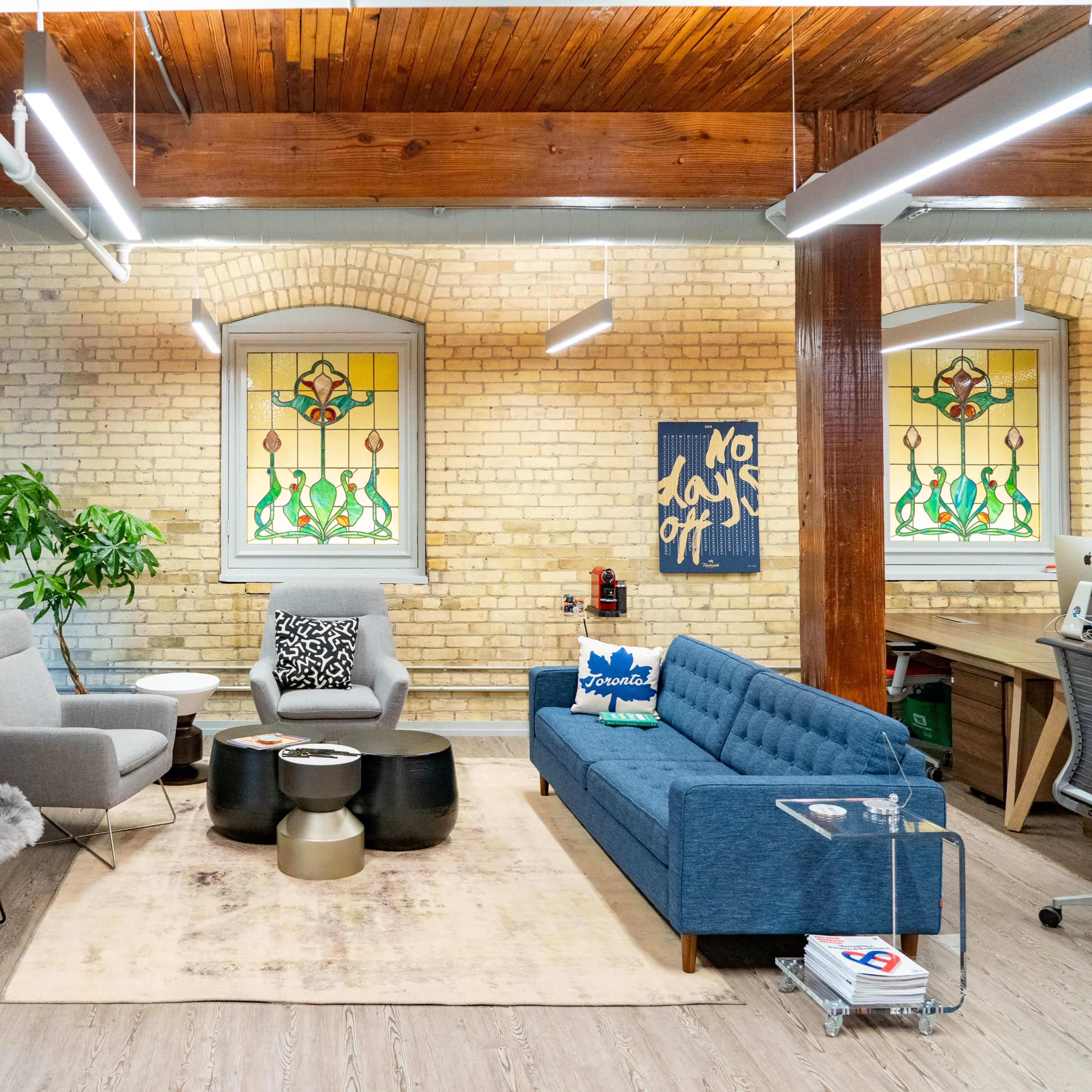 A modern office space featuring a seating area with a blue couch, gray chairs, a coffee table, and large windows with decorative stained glass.