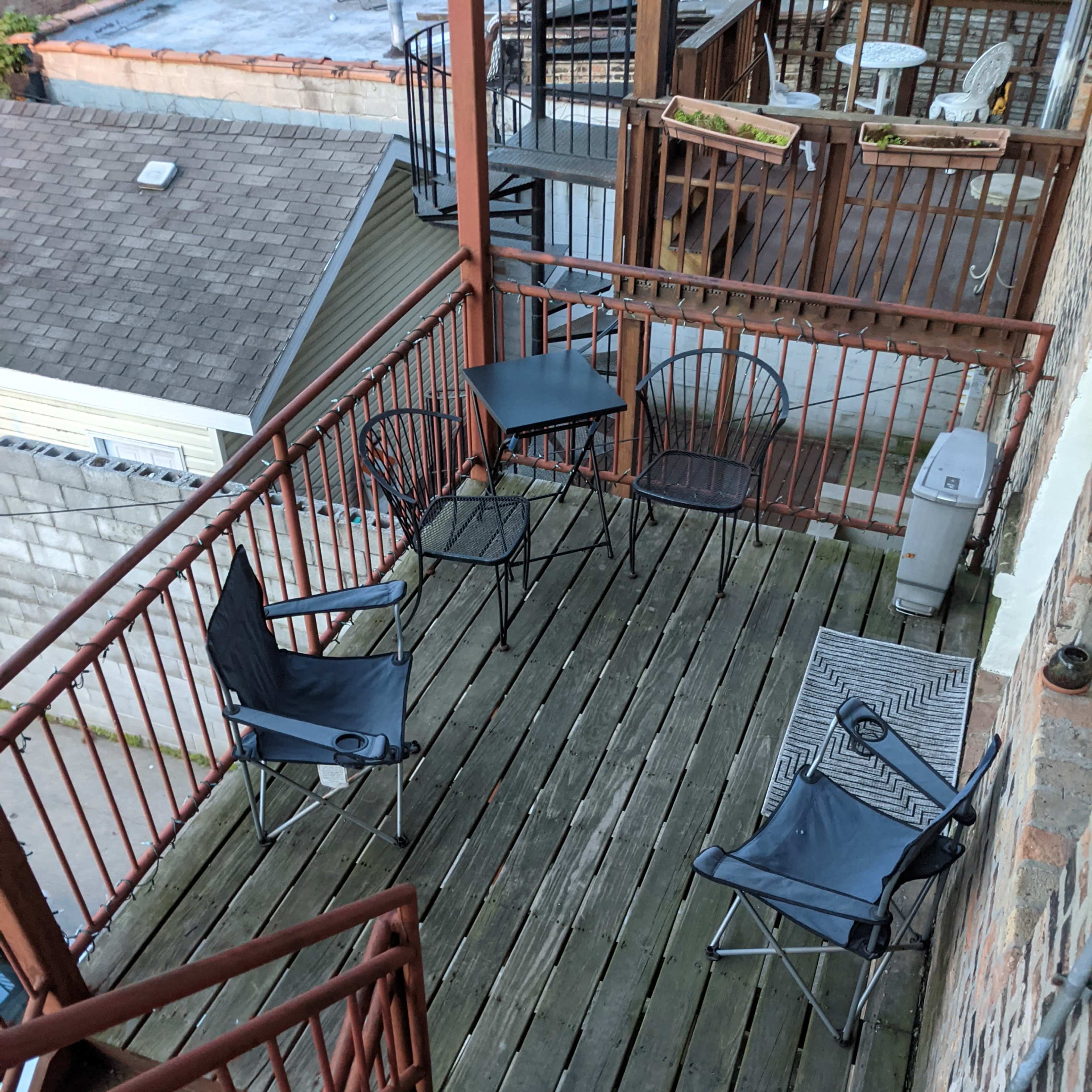 A small outdoor seating area with a black table and four folding chairs is located on a wooden deck, surrounded by a railing.