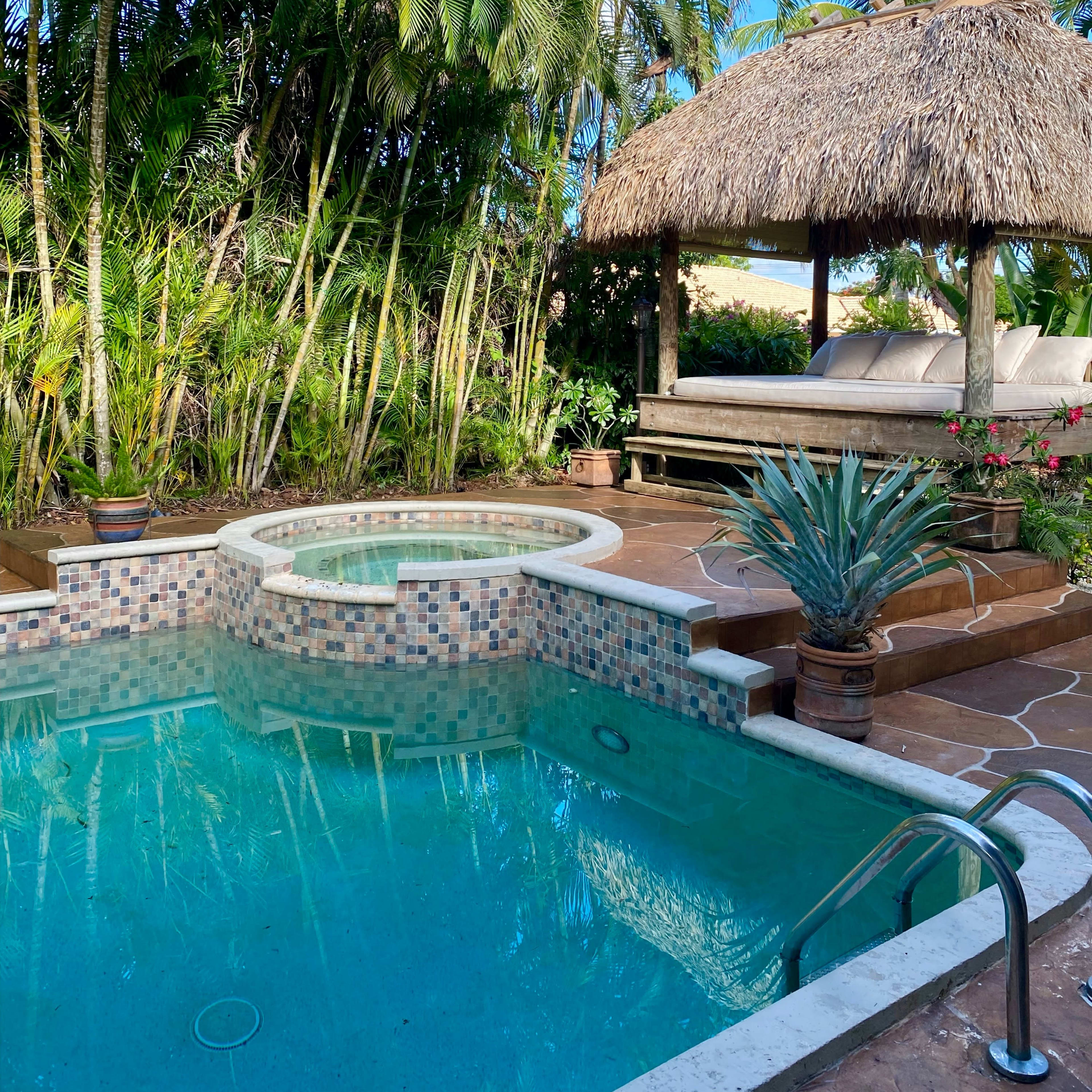 A sparkling hot tub is surrounded by lush palm trees and a thatched-roof gazebo with a couch.