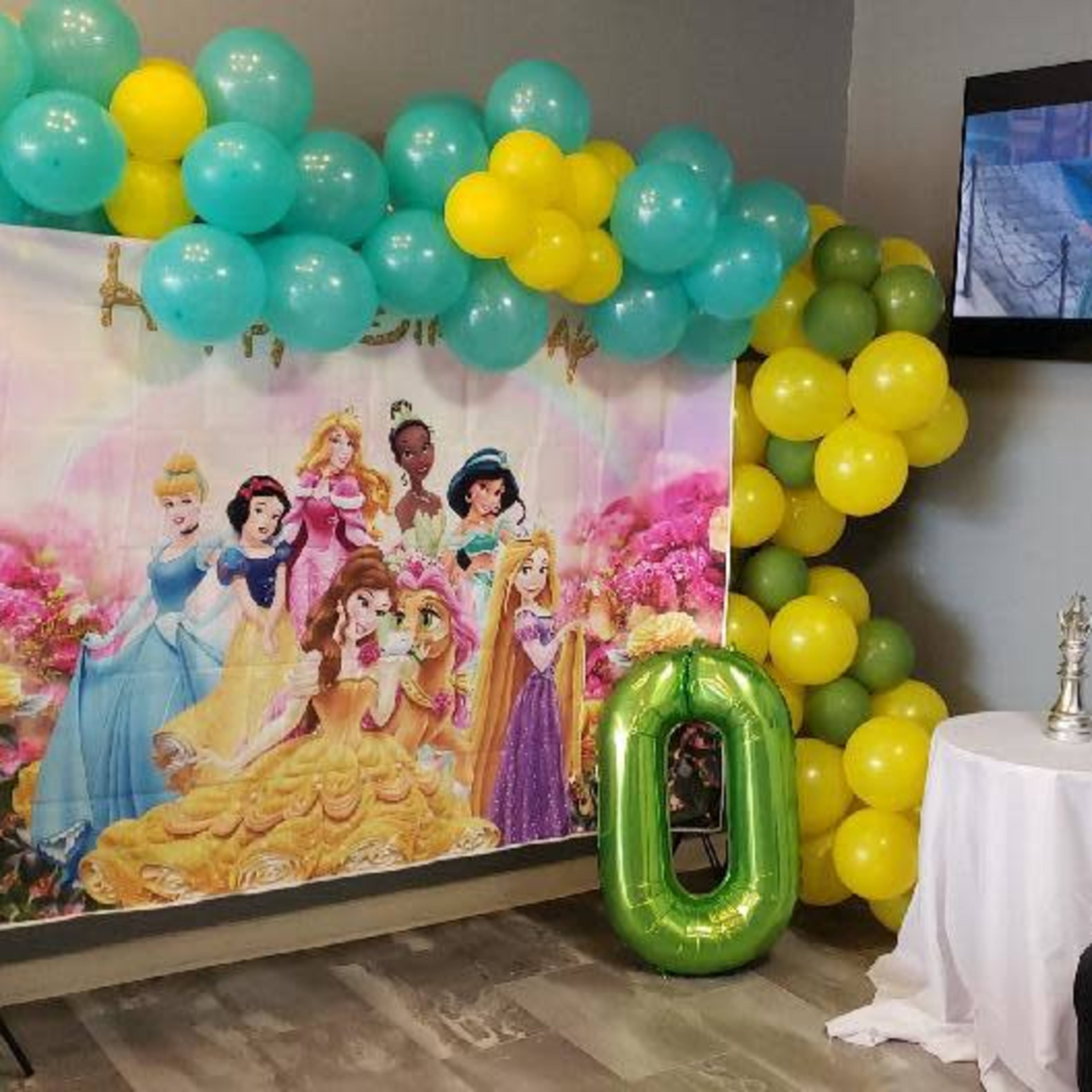 A decorated corner features a colorful balloon backdrop with a princess theme, a table with a white cloth, a plush black chair, and a large "10" balloon.