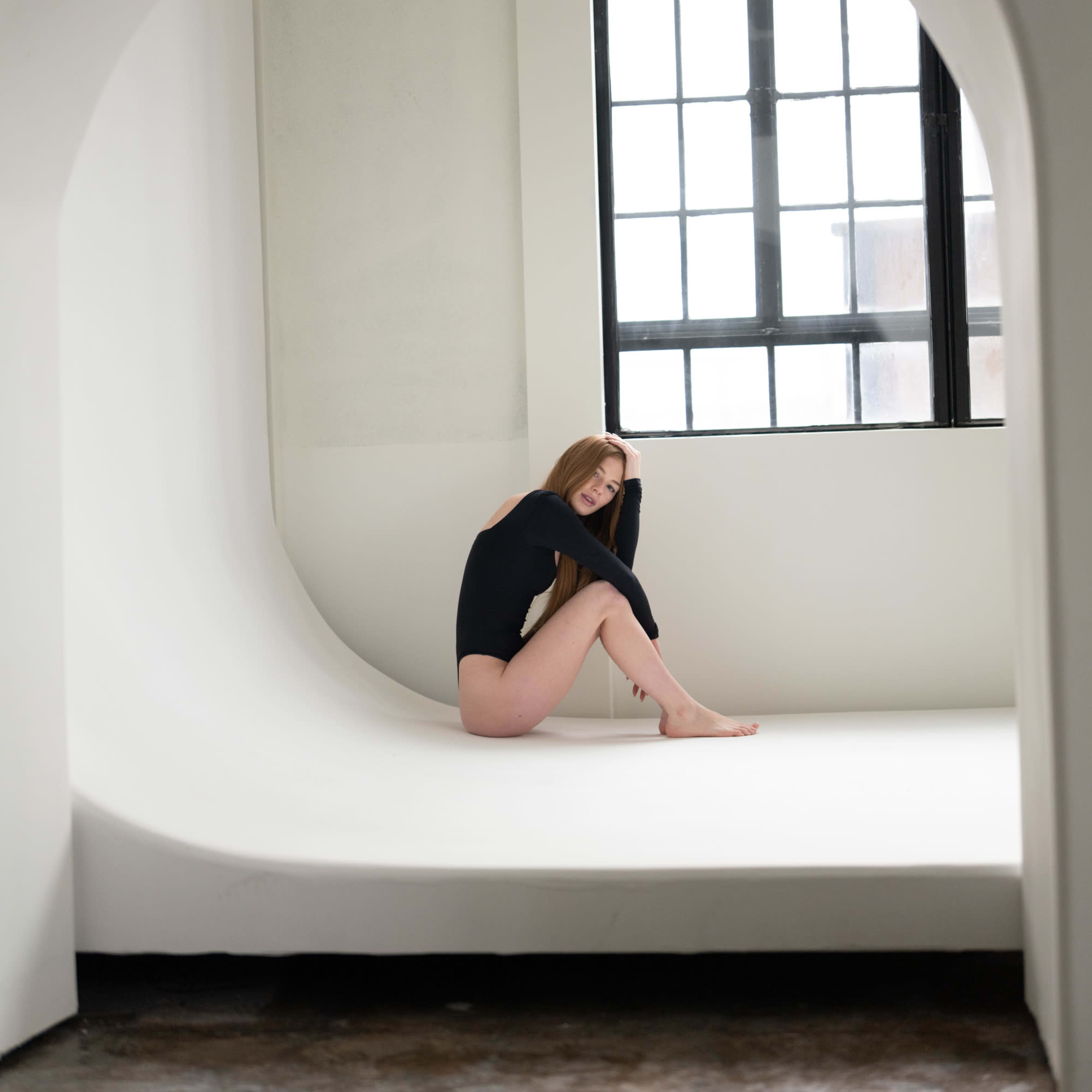 A person sits in a minimalist, curved white space near a large window with black frames.