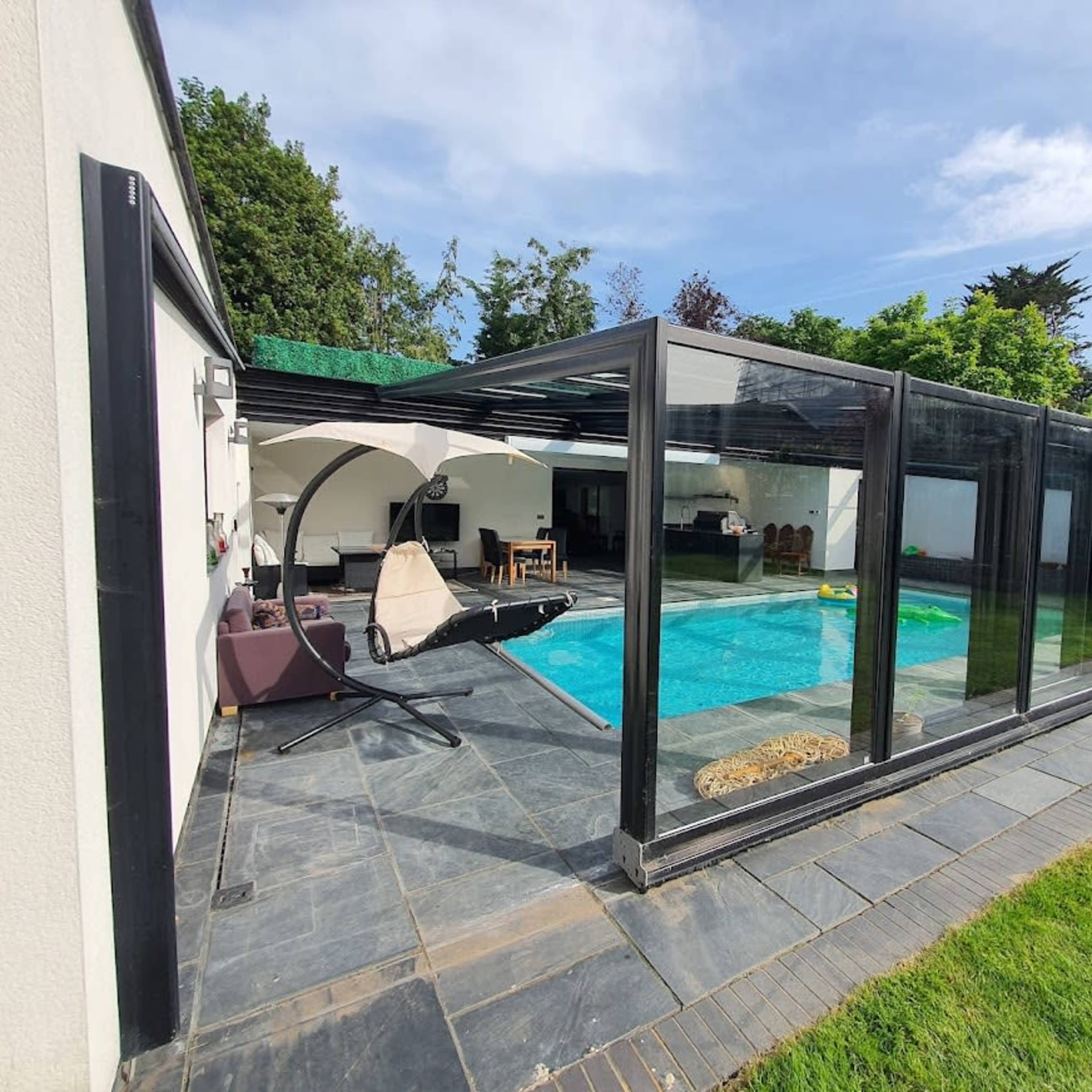 A modern pool area enclosed with glass panels, featuring a swimming pool surrounded by stone tiles and a grassy lawn.