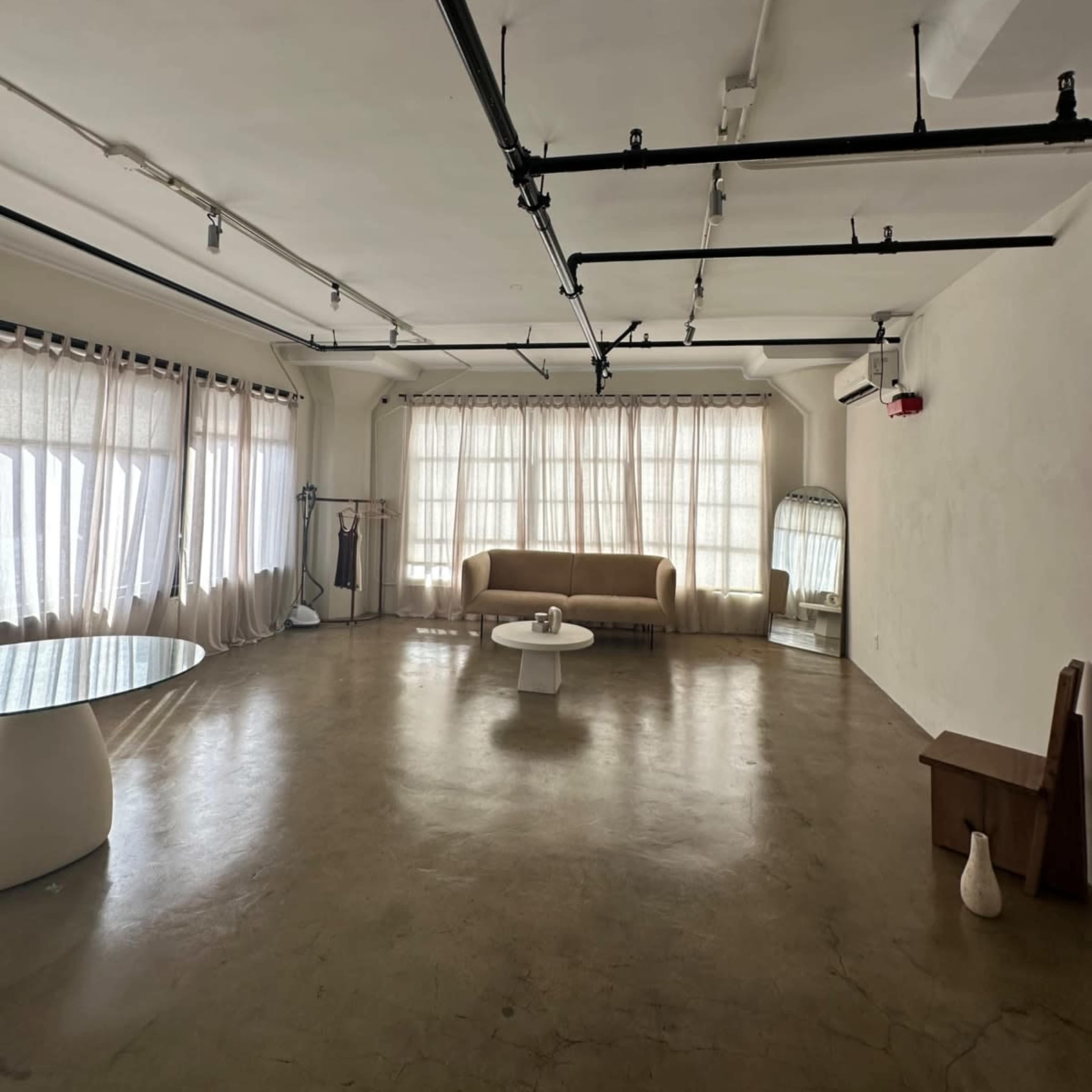 An empty, modern room with large windows covered by light curtains, a beige sofa, a white coffee table, and polished concrete flooring.