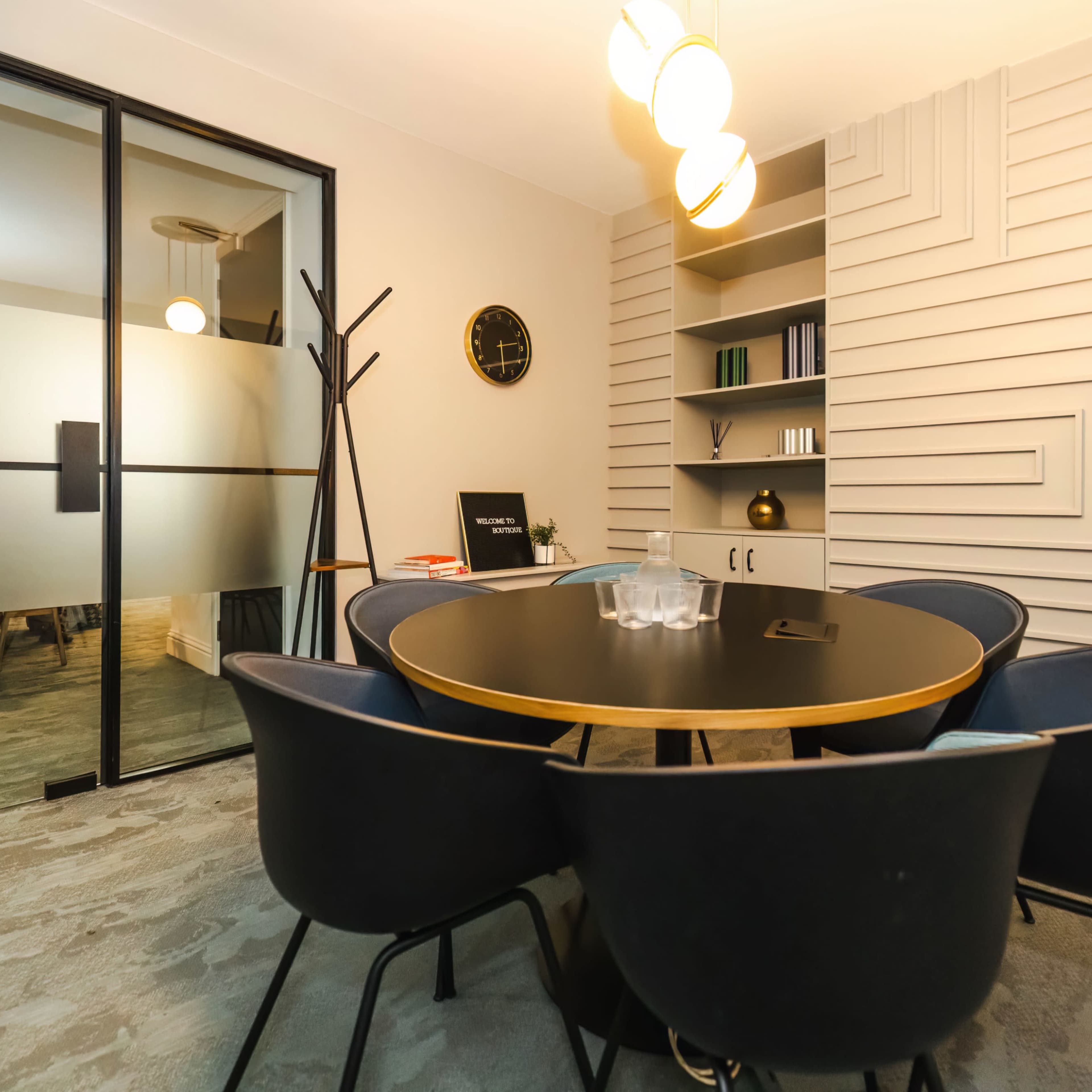 A modern conference room features a round black table surrounded by four blue chairs, with a glass partition and shelves in the background.