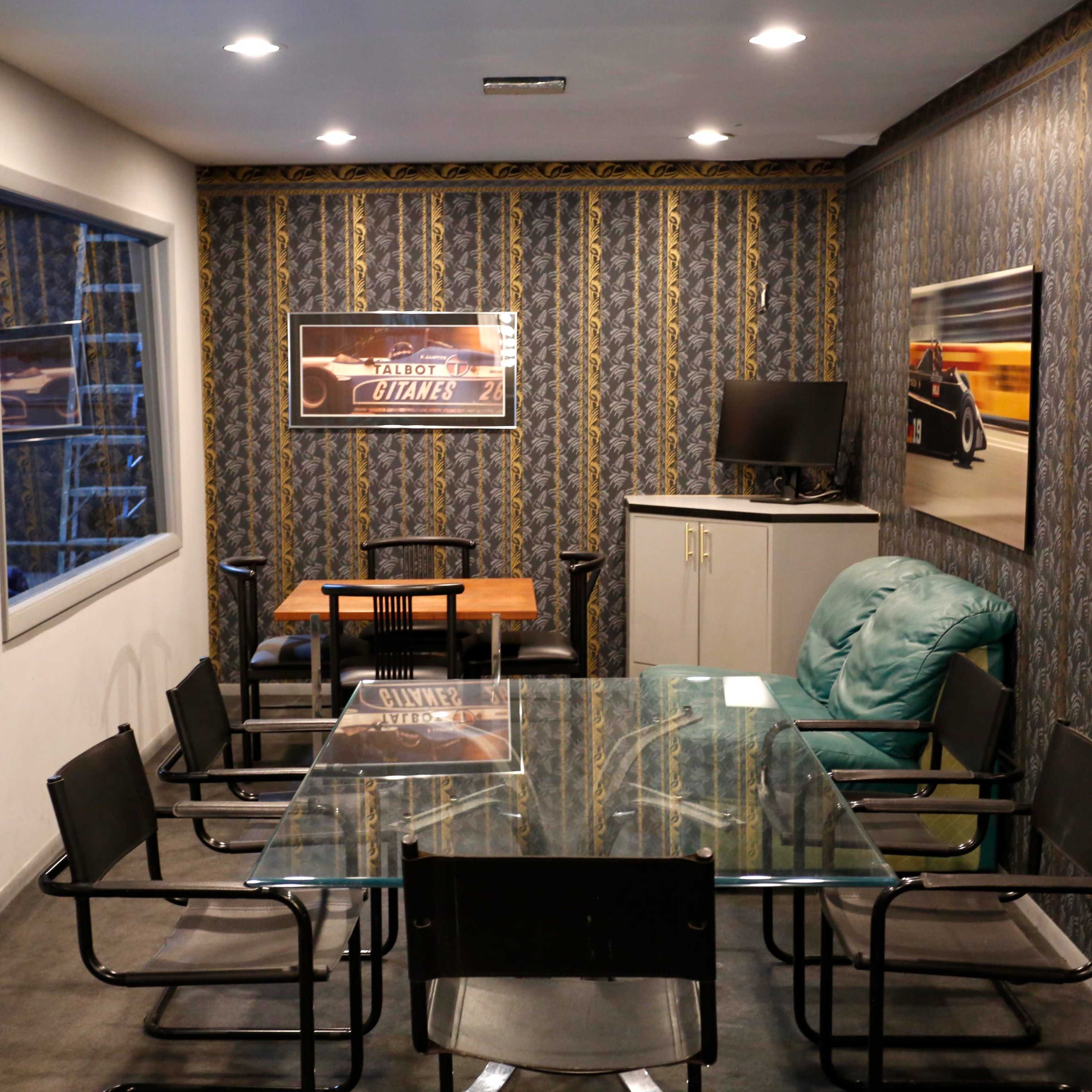 A small meeting room with a glass table, black chairs, a wooden table, a green armchair, and framed posters on the patterned walls.