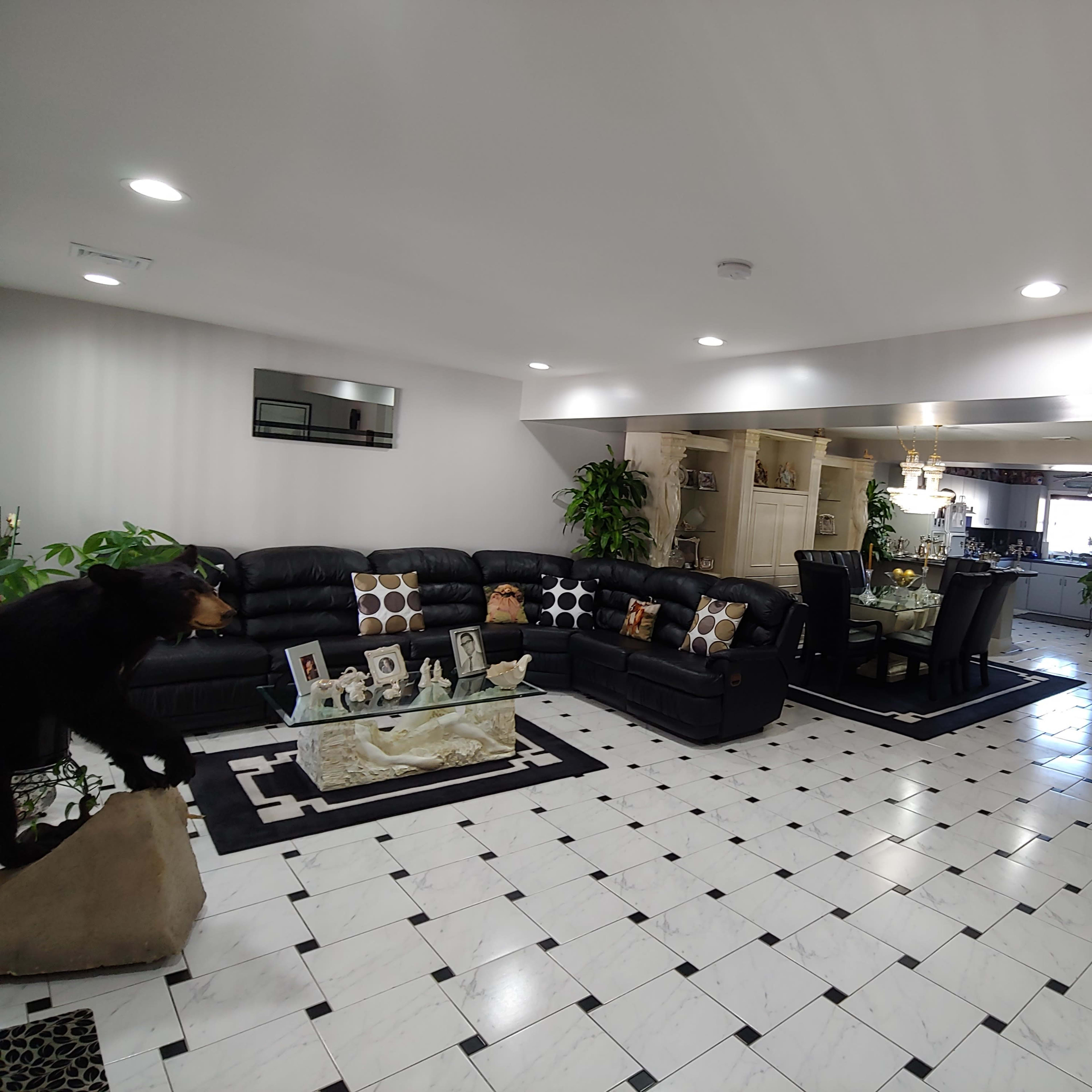 A living room with black leather furniture, a glass coffee table, and a dining area in the background, featuring a tiled floor with black and white patterns.