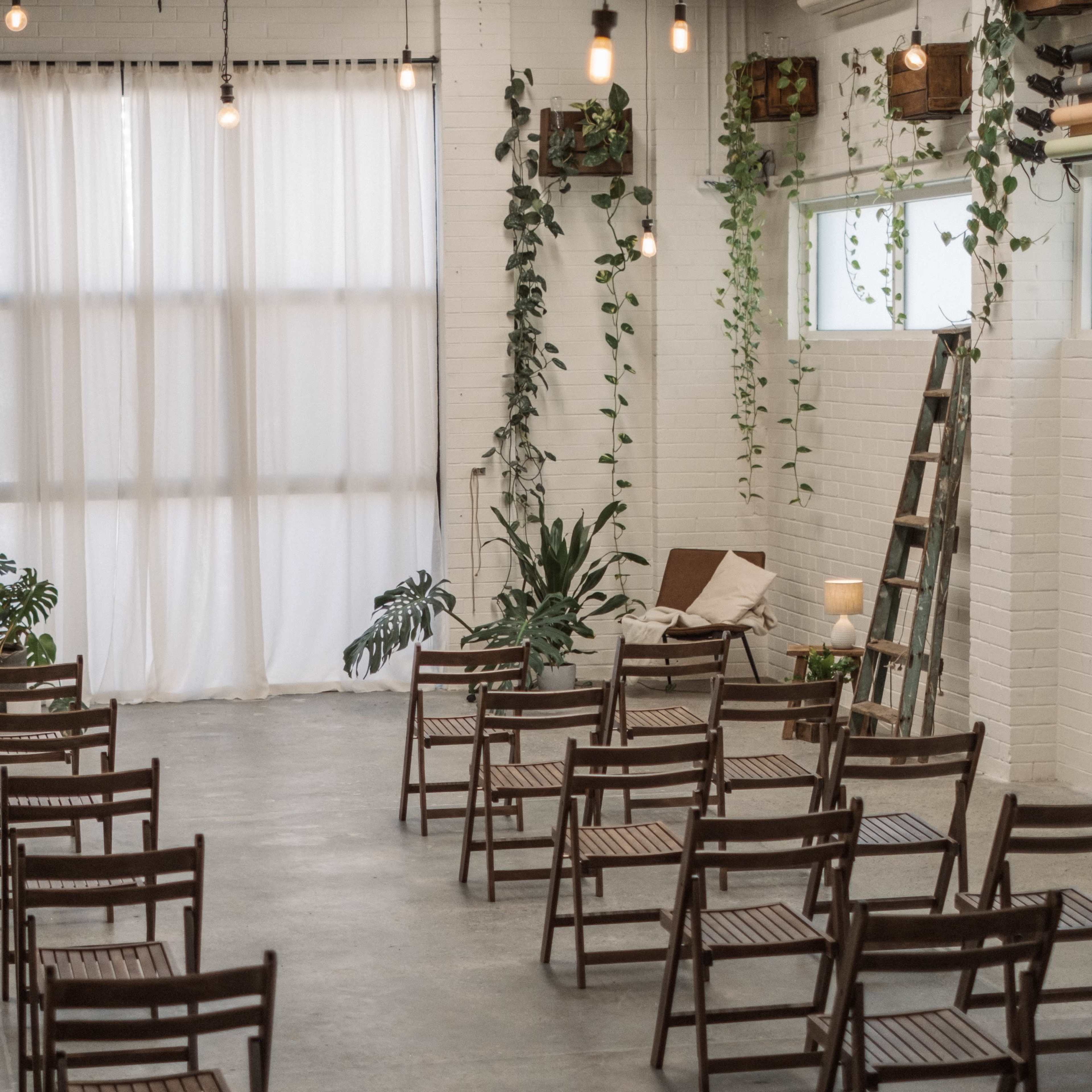 A spacious indoor venue with wooden chairs arranged for an event and tall white curtains, accented by plants and light fixtures.