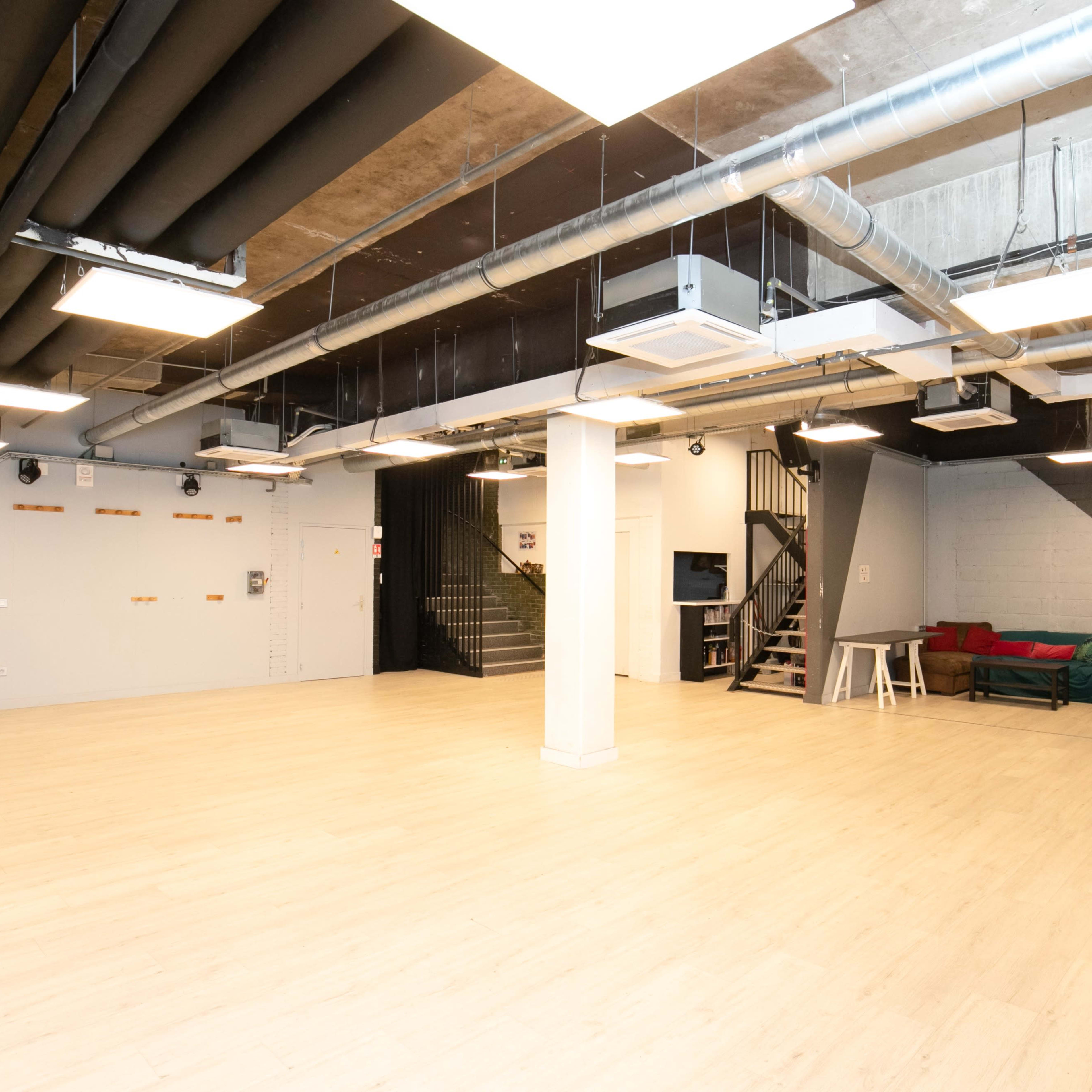 A spacious, empty room with light wooden flooring and industrial-style ceilings, featuring a staircase in the back and a small couch against the wall.