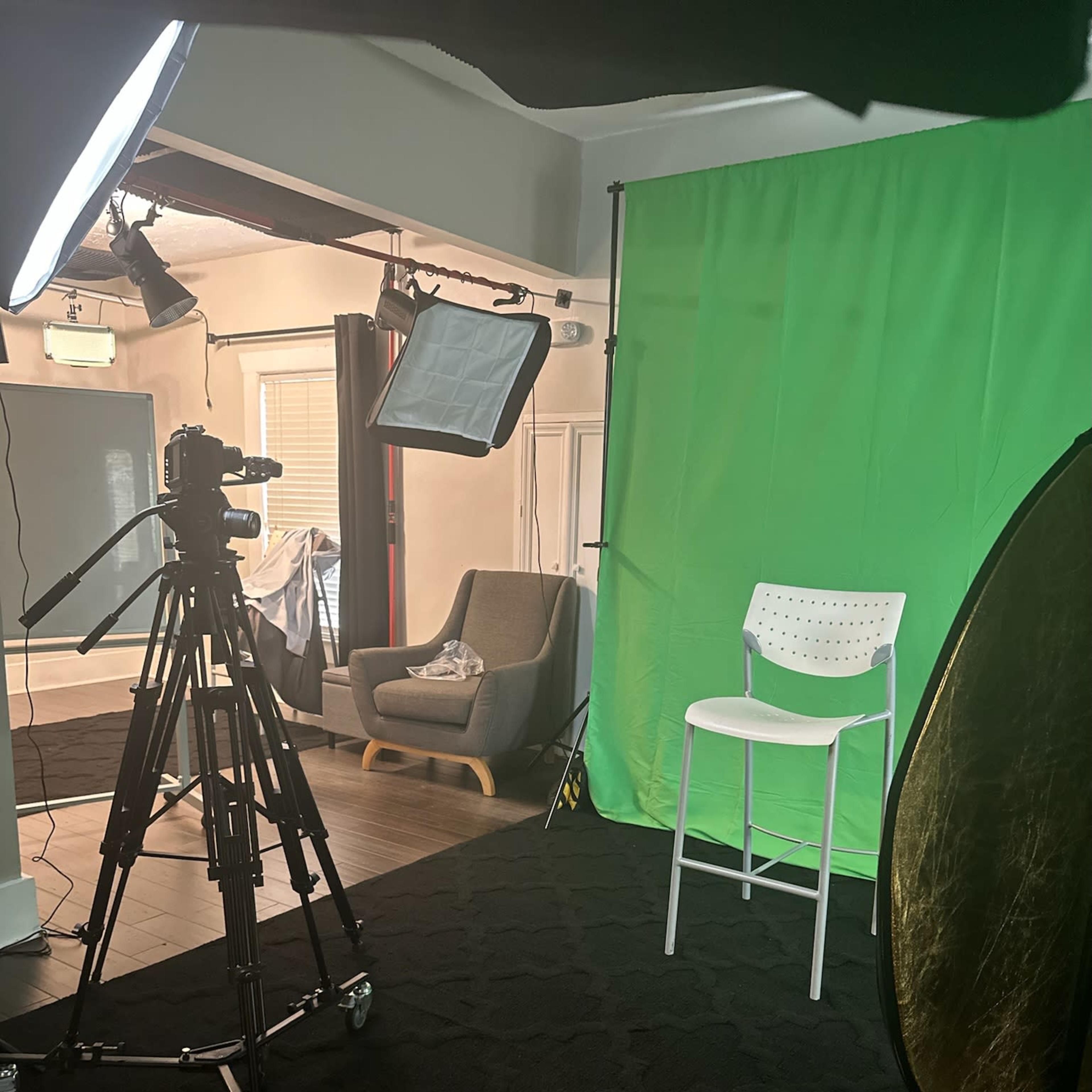 A video recording setup featuring a camera on a tripod aimed at a white chair in front of a green screen, with studio lights positioned to the side.