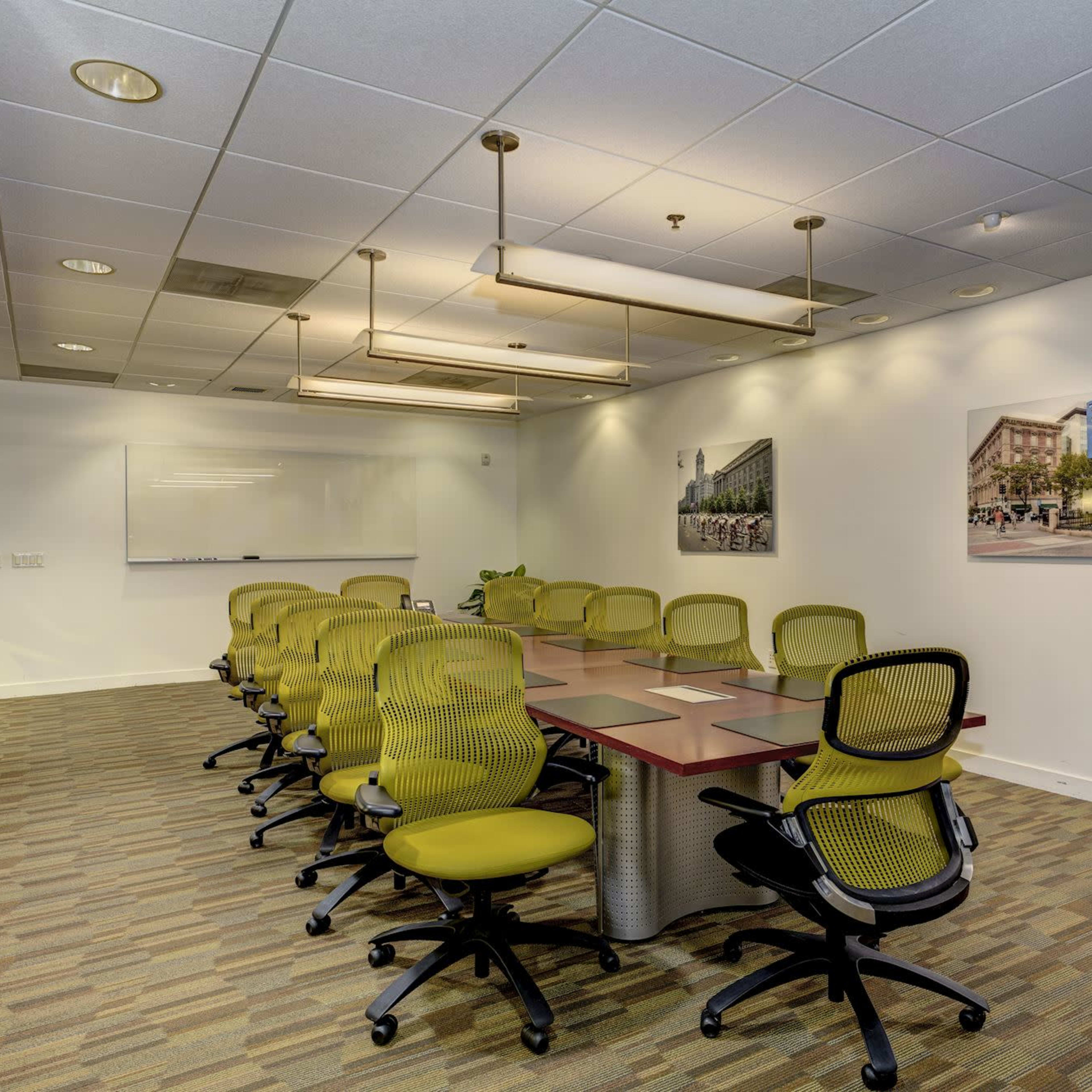 A modern conference room features a long table surrounded by green ergonomic chairs, with artwork on the walls and a whiteboard at one end.