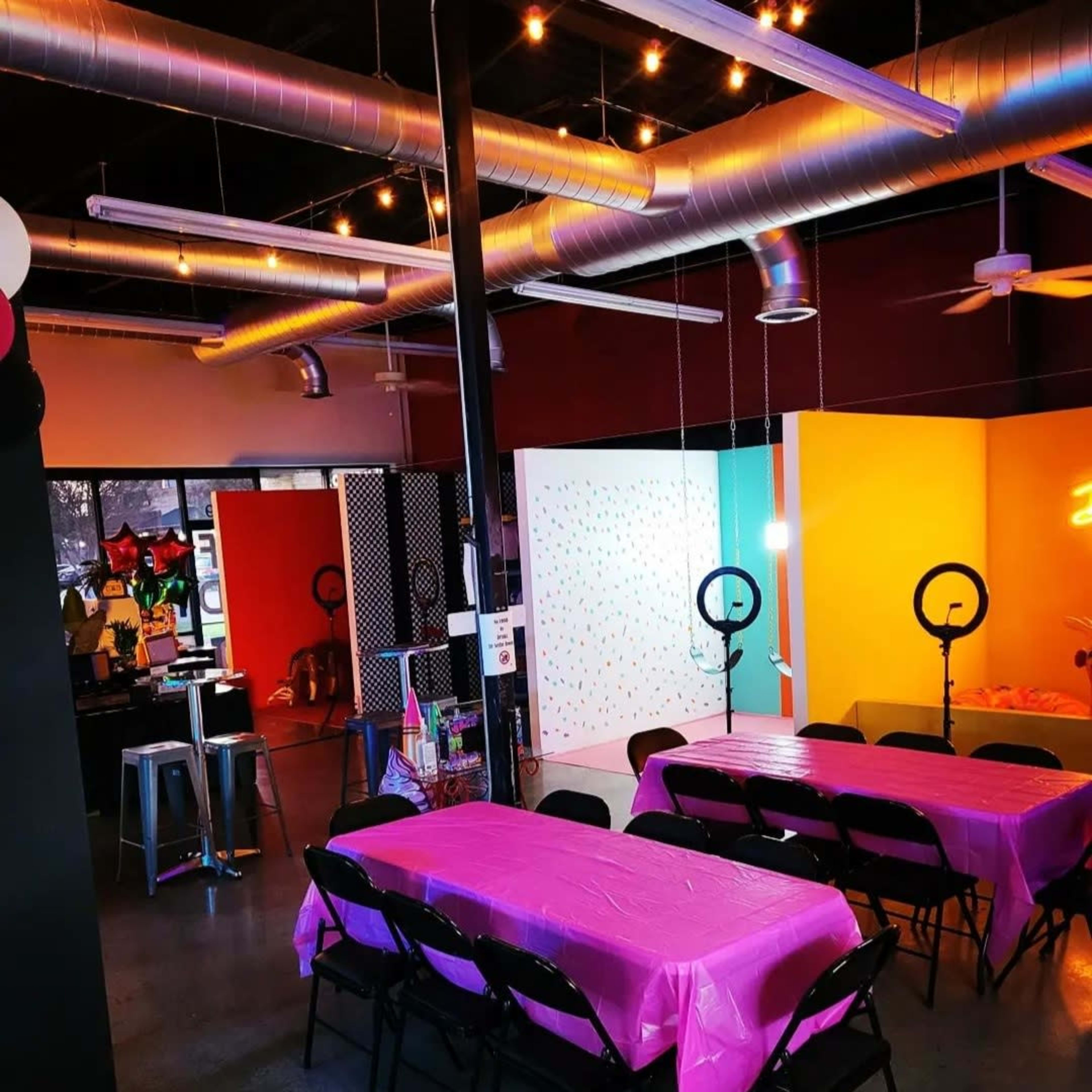 A party venue with several pink tables, bright colored walls, and ring lights set up.