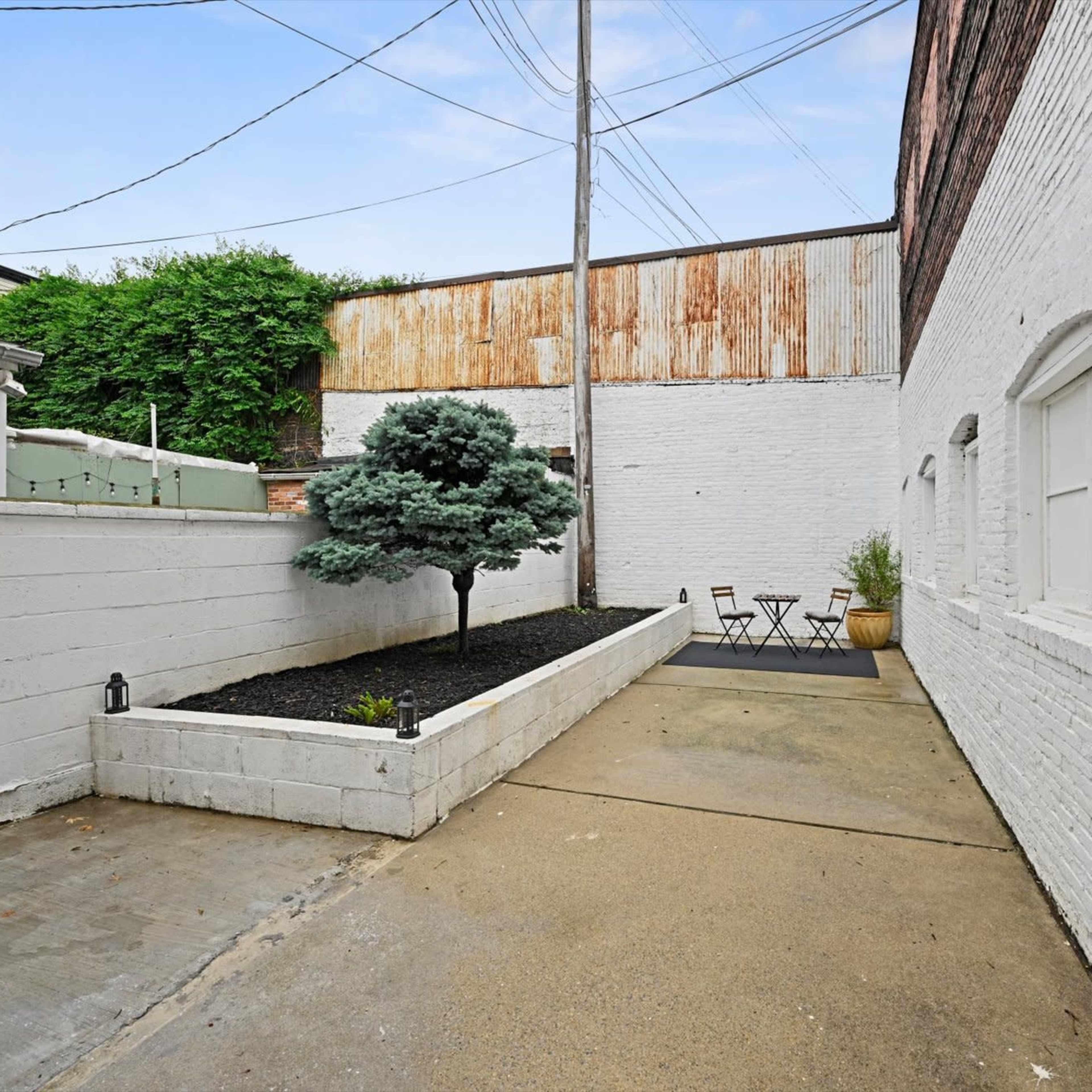 A narrow outdoor space with a gravel area, a small tree, and a seating arrangement against a white brick wall.