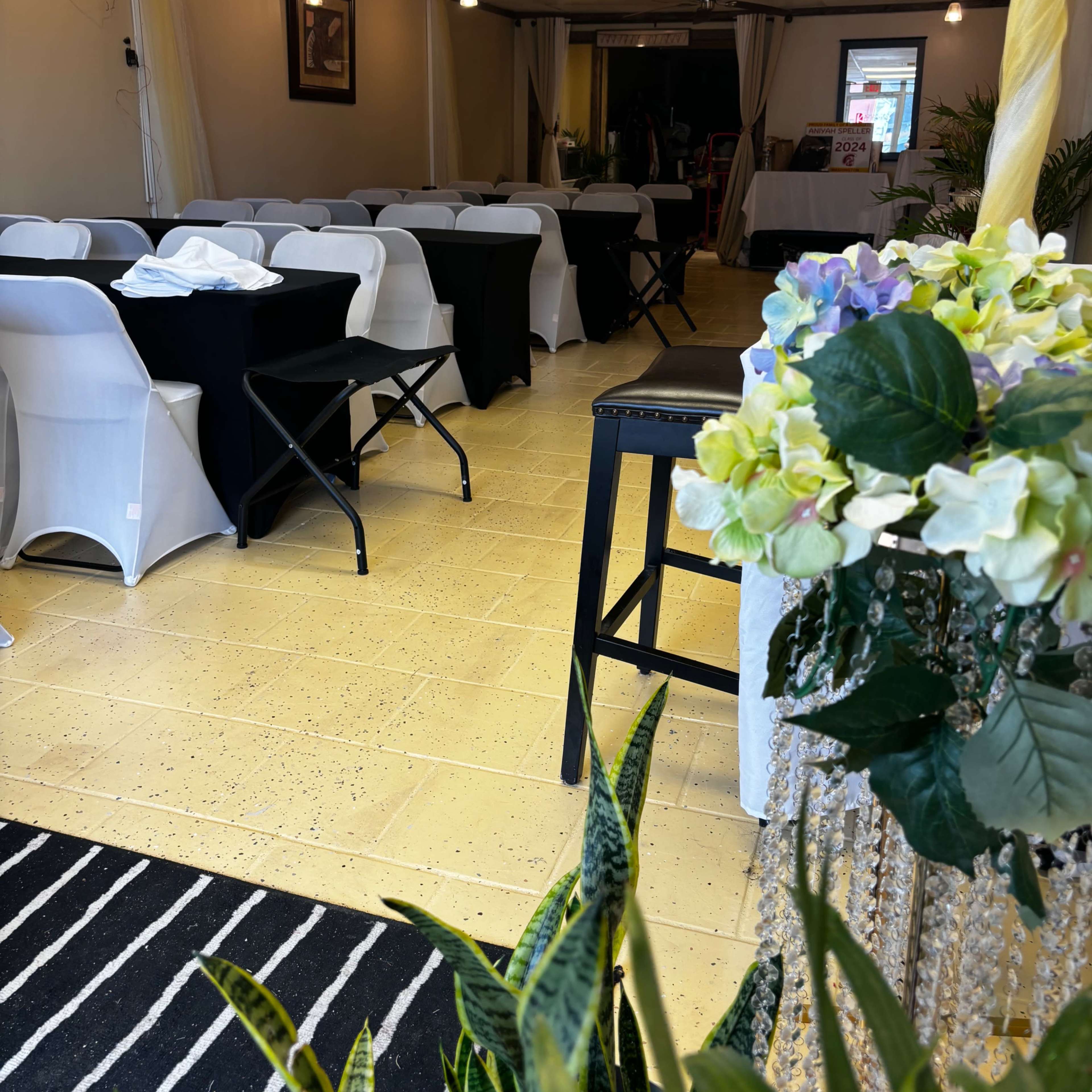 A partially decorated indoor event space with tables covered in black and white linens, a striped rug, and plants arranged in the foreground.