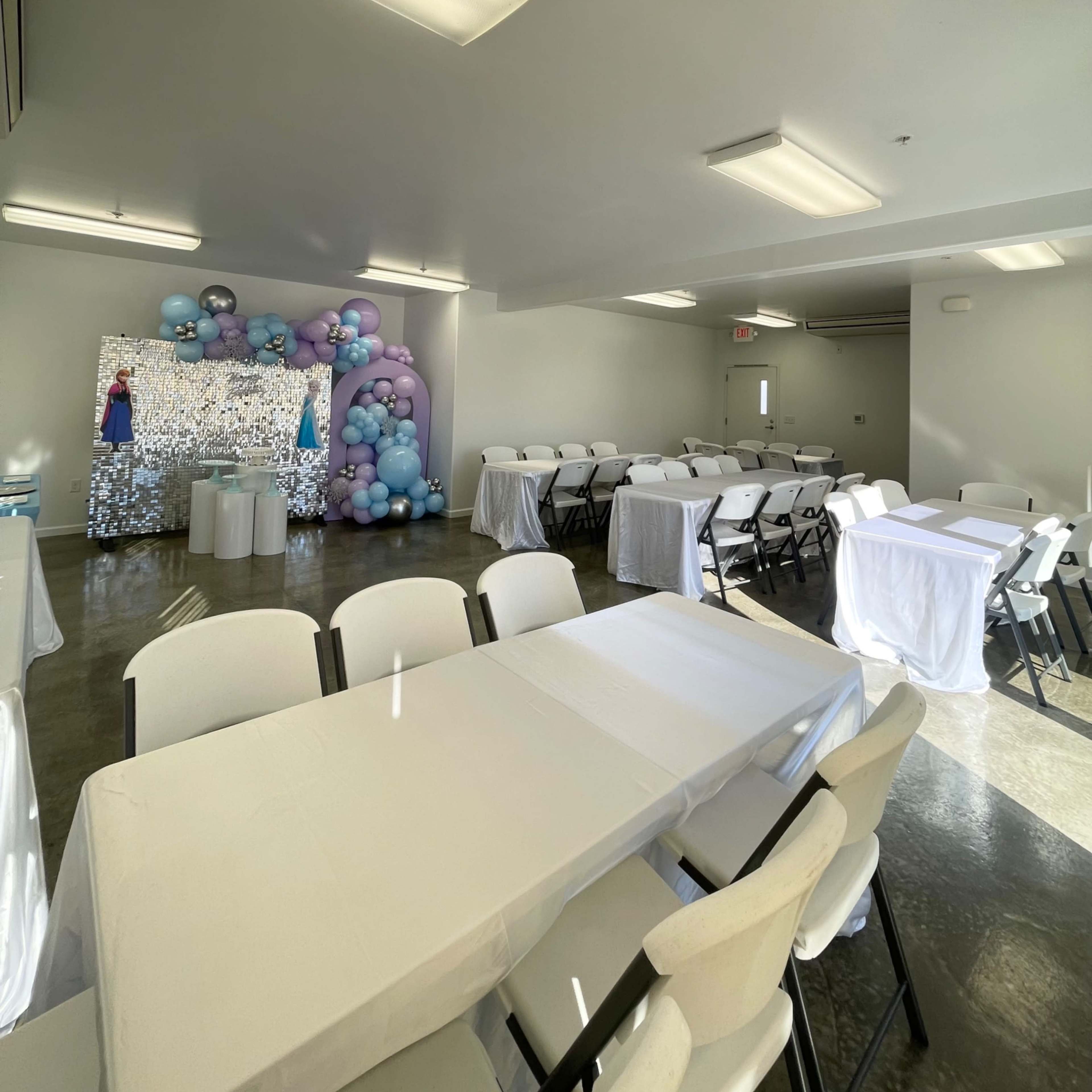 A spacious event room with white tables and chairs set up, featuring a large balloon arch and a decorative backdrop at one end.