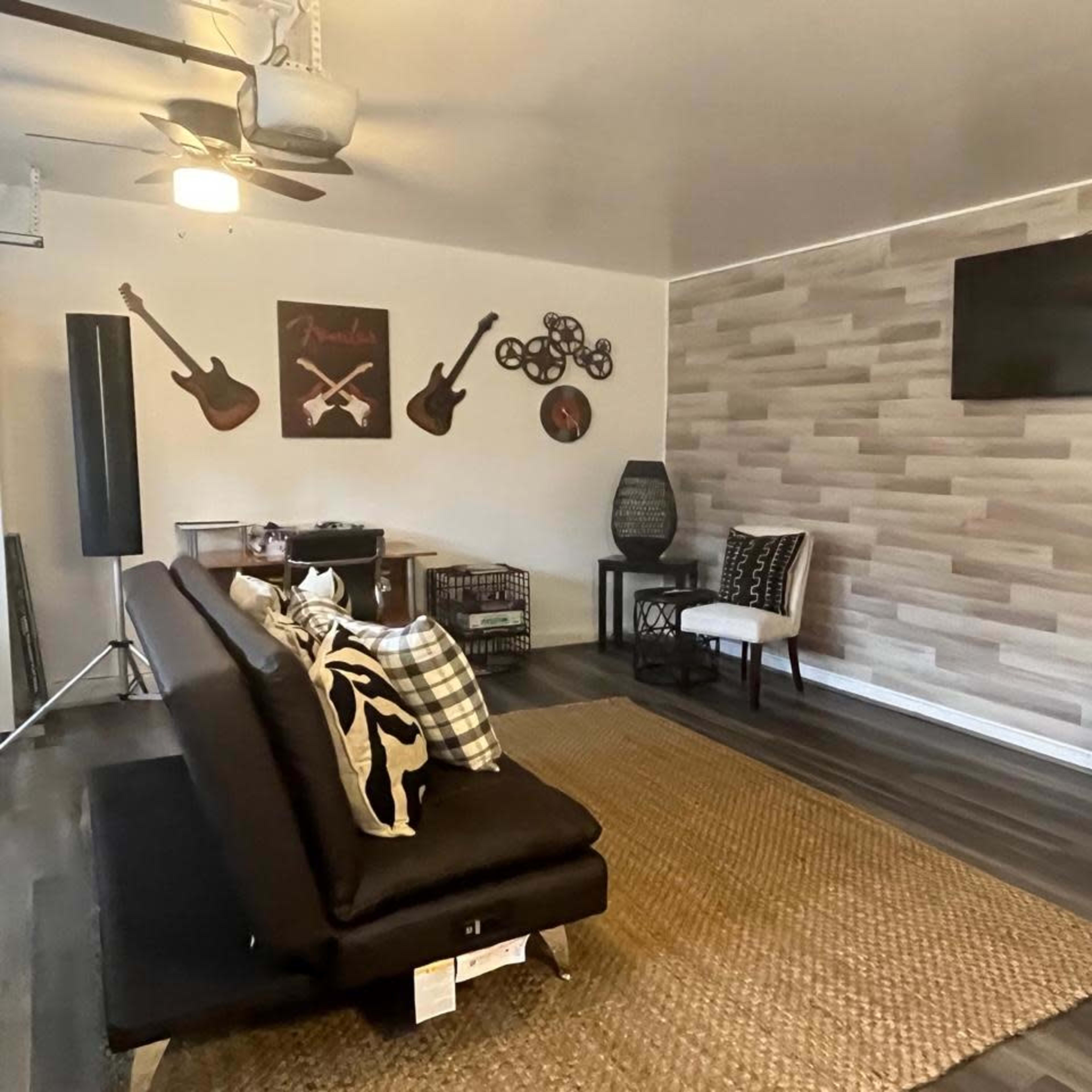 A modern living room featuring a black sofa, a wall-mounted television, a patterned area rug, and decorative items such as guitars on the wall and various seating options.