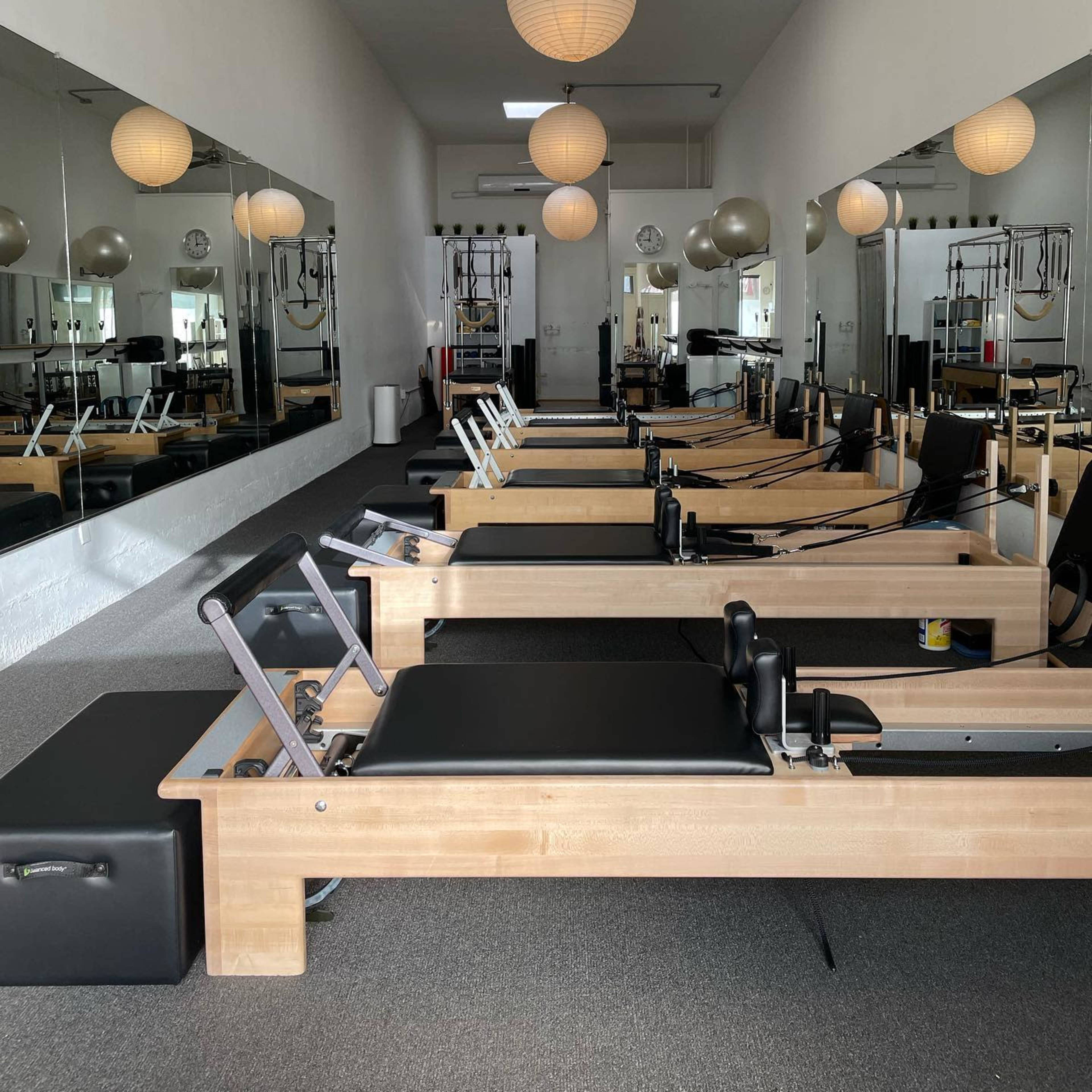A well-lit Pilates studio featuring multiple reformer machines aligned in rows, with large mirrors reflecting the space and pendant lights hanging from the ceiling.