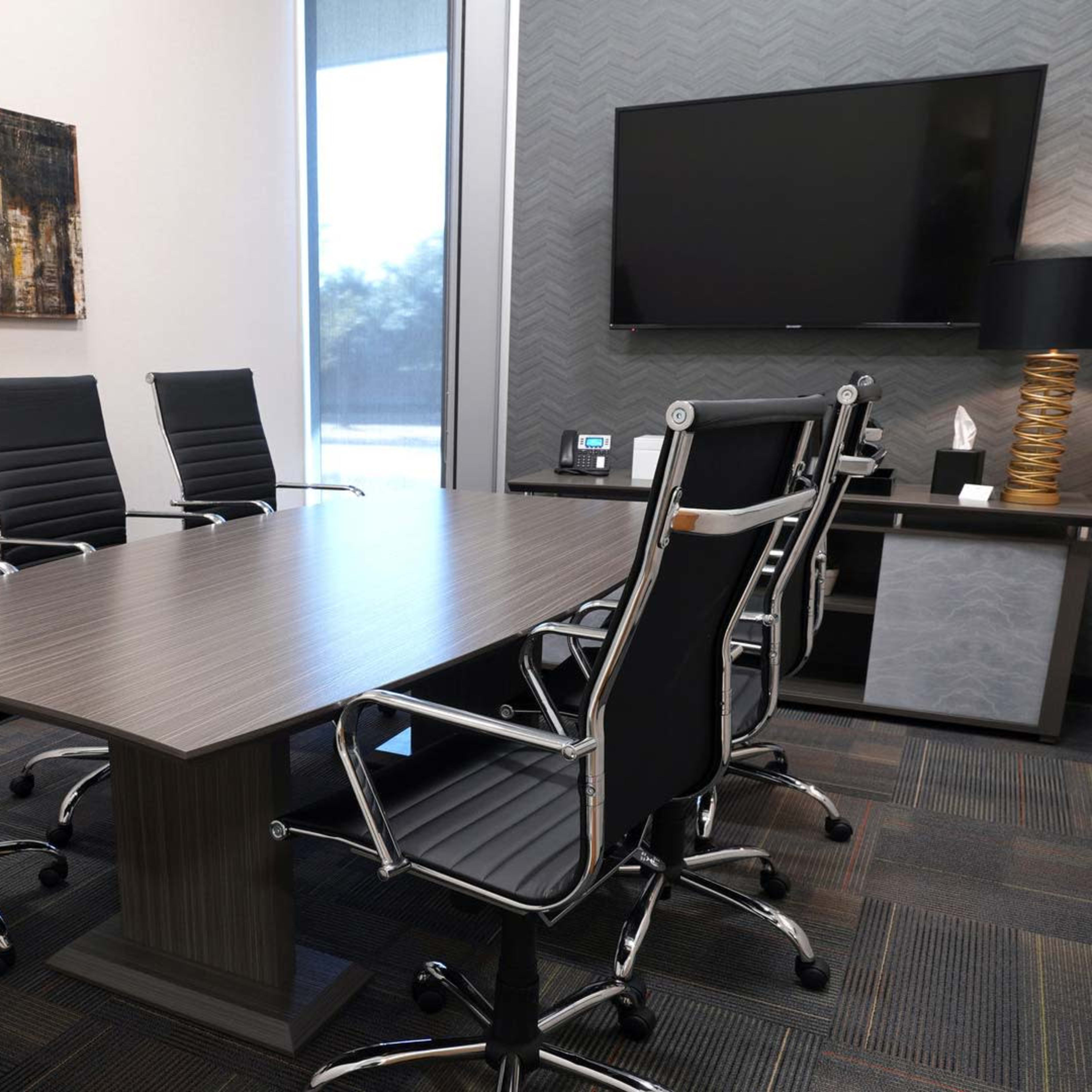 A modern conference room features a large rectangular table surrounded by six black ergonomic chairs, with a television mounted on the wall and a decorative painting in the background.