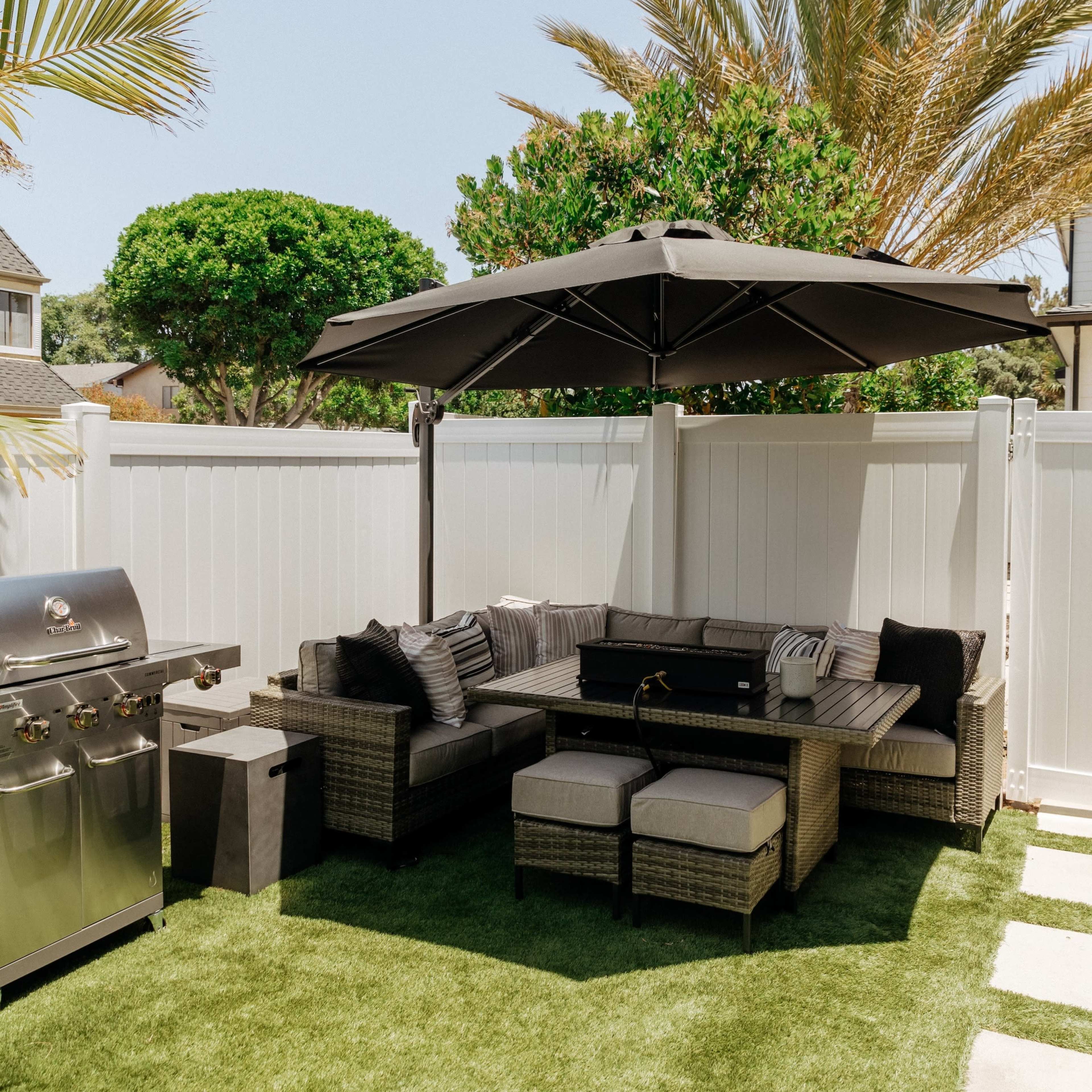 An outdoor seating area with a sectional sofa under a large umbrella, surrounded by palm trees and a grill on the side.