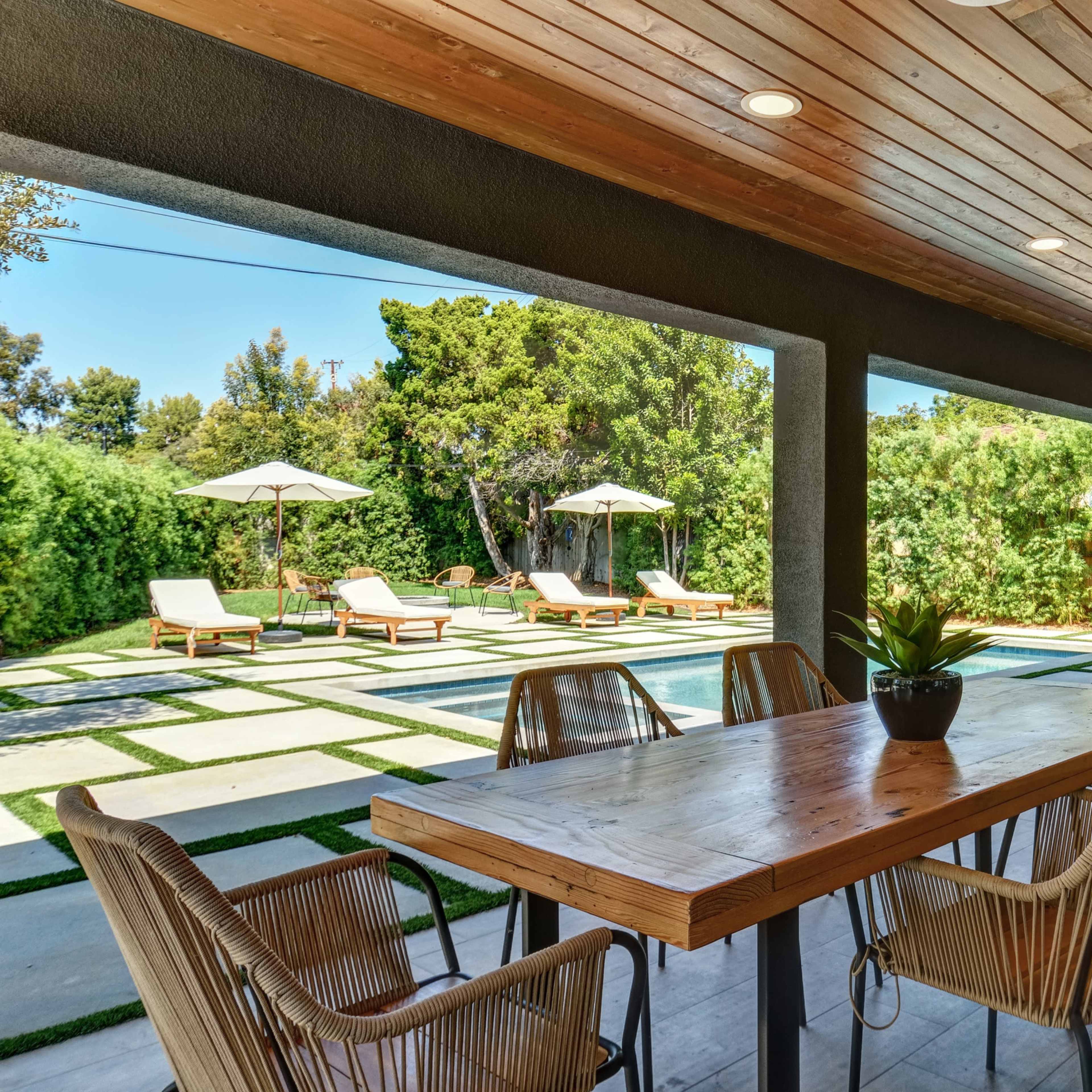 A covered patio features a large wooden dining table surrounded by chairs, with a view of a landscaped yard containing lounge chairs and umbrellas.