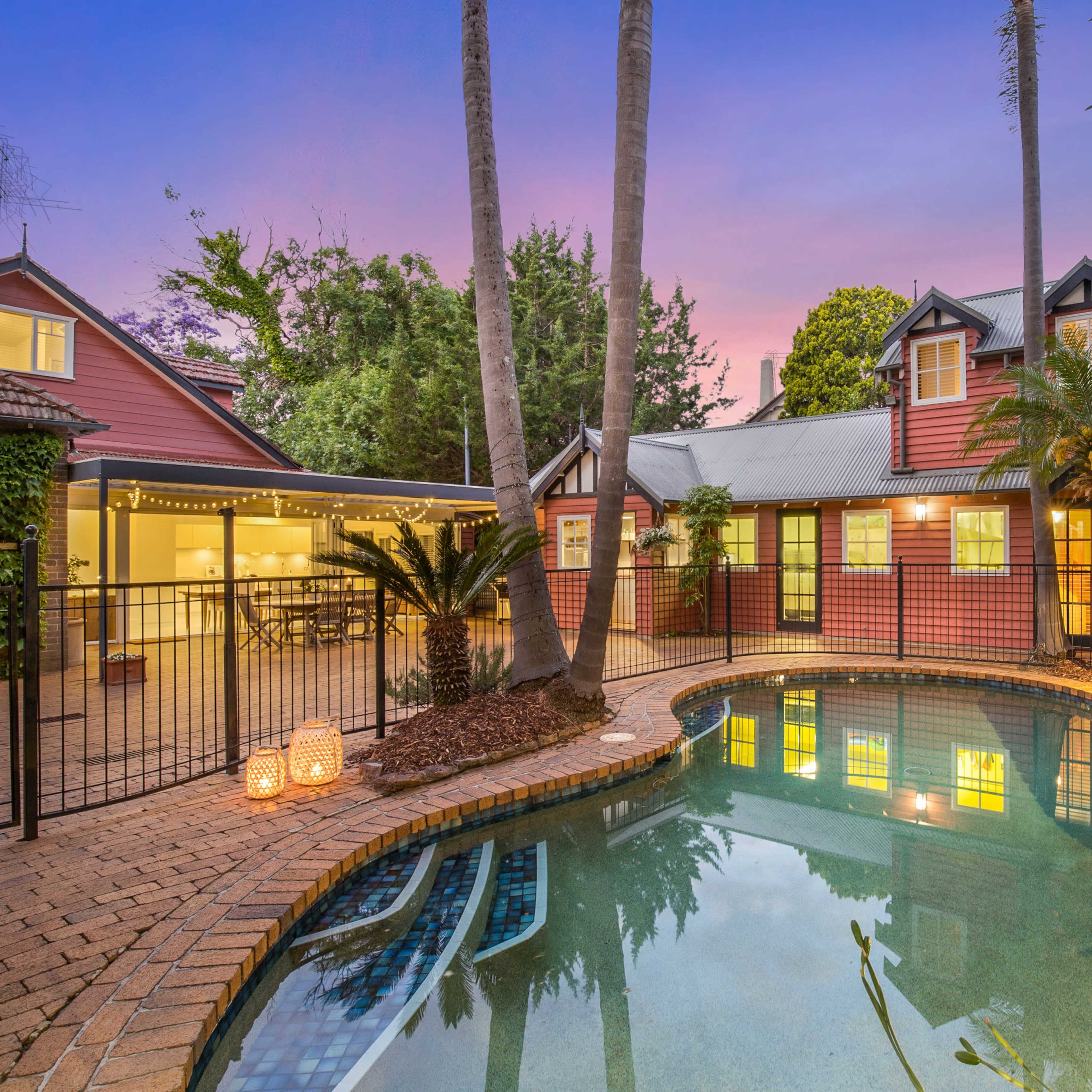 A backyard featuring a swimming pool surrounded by a red and white house, palm trees, and a dining area with outdoor seating at dusk.