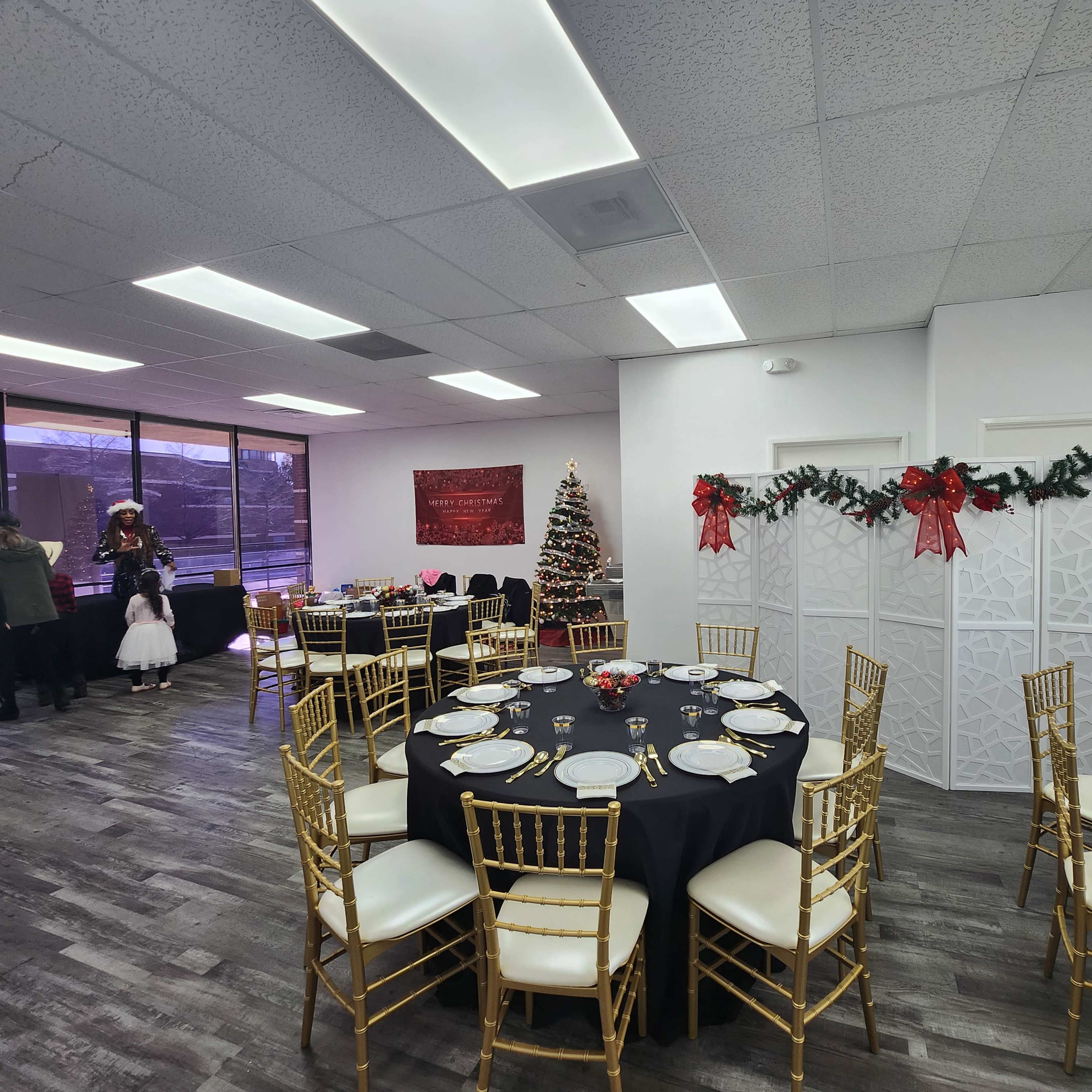 A festive event space decorated with a Christmas tree, tables set with gold and black tableware, and holiday decorations on the walls.