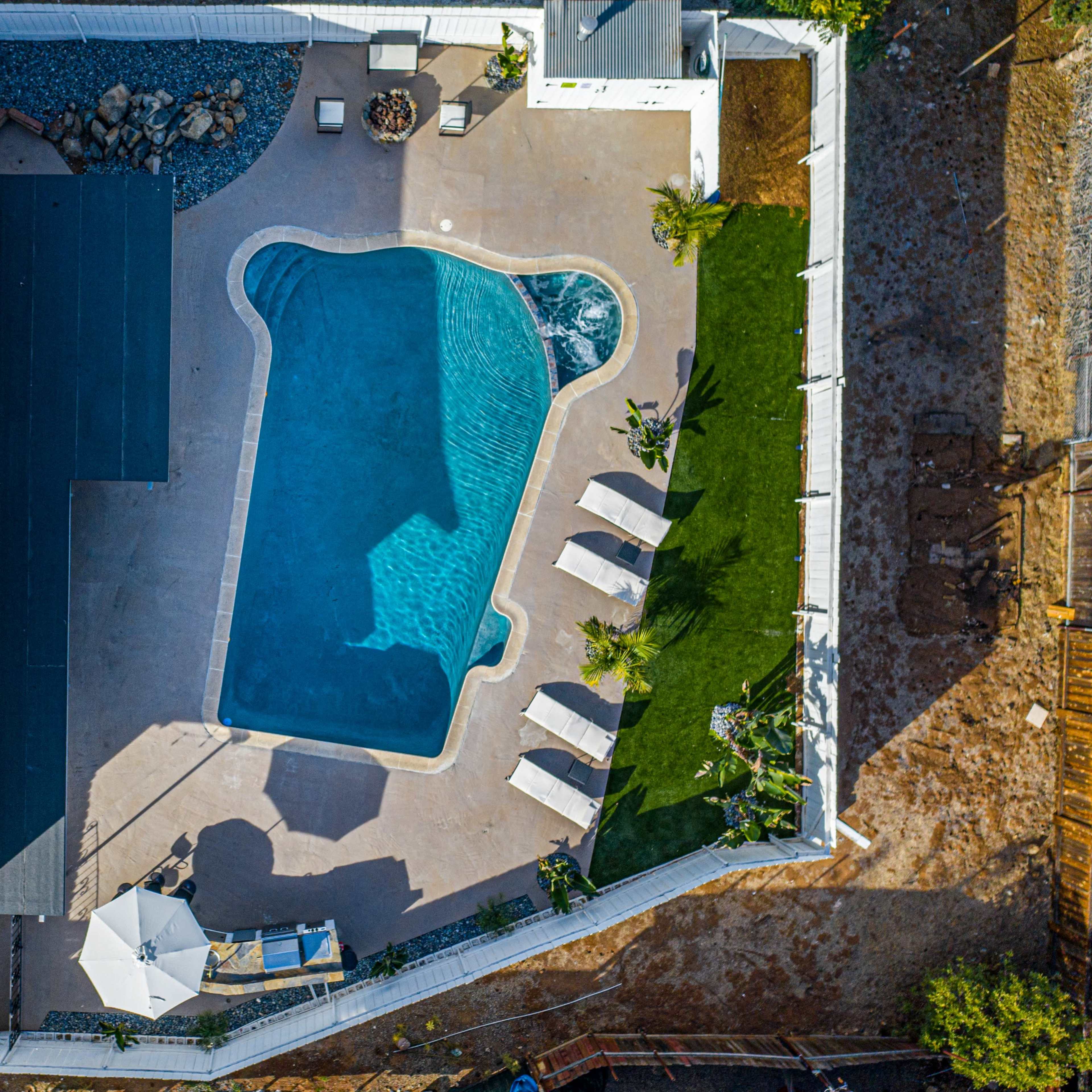 An aerial view of a backyard featuring a swimming pool surrounded by lounge chairs, structured landscaping, and a covered patio area.