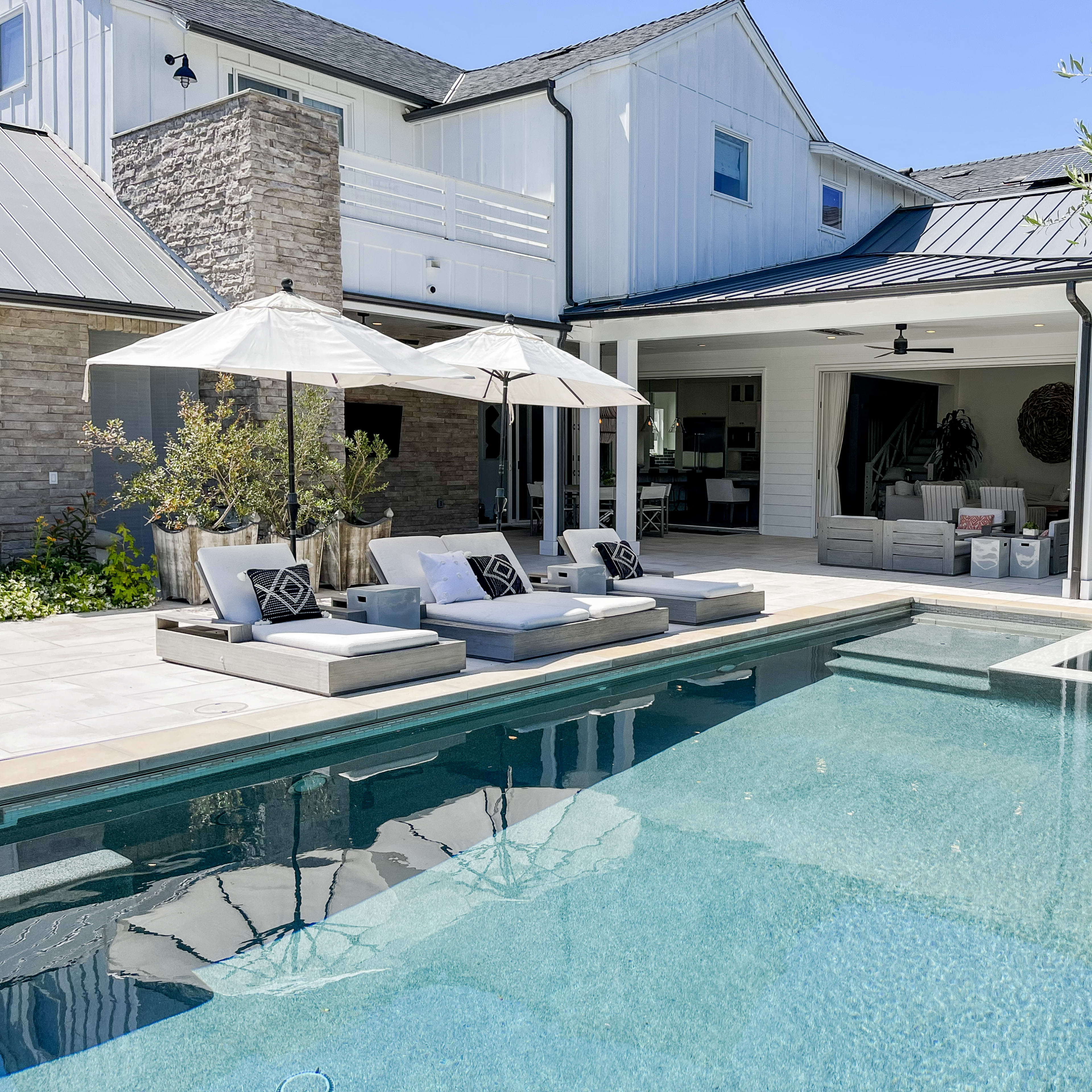 A swimming pool with lounge chairs and umbrellas is situated in a patio area adjacent to a modern house with a covered outdoor living space.