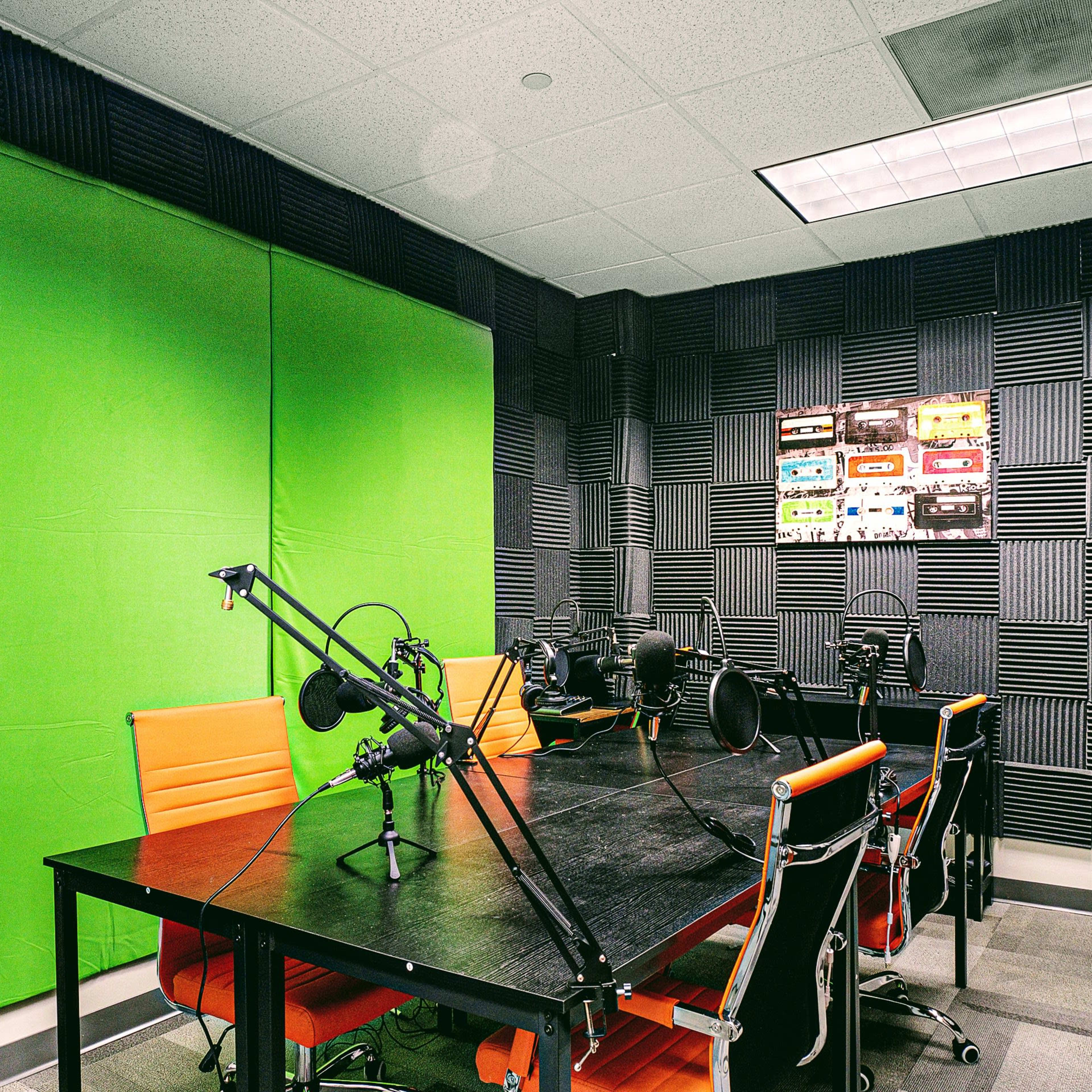A soundproof recording studio with a green backdrop, a large table, multiple microphones, and orange chairs.