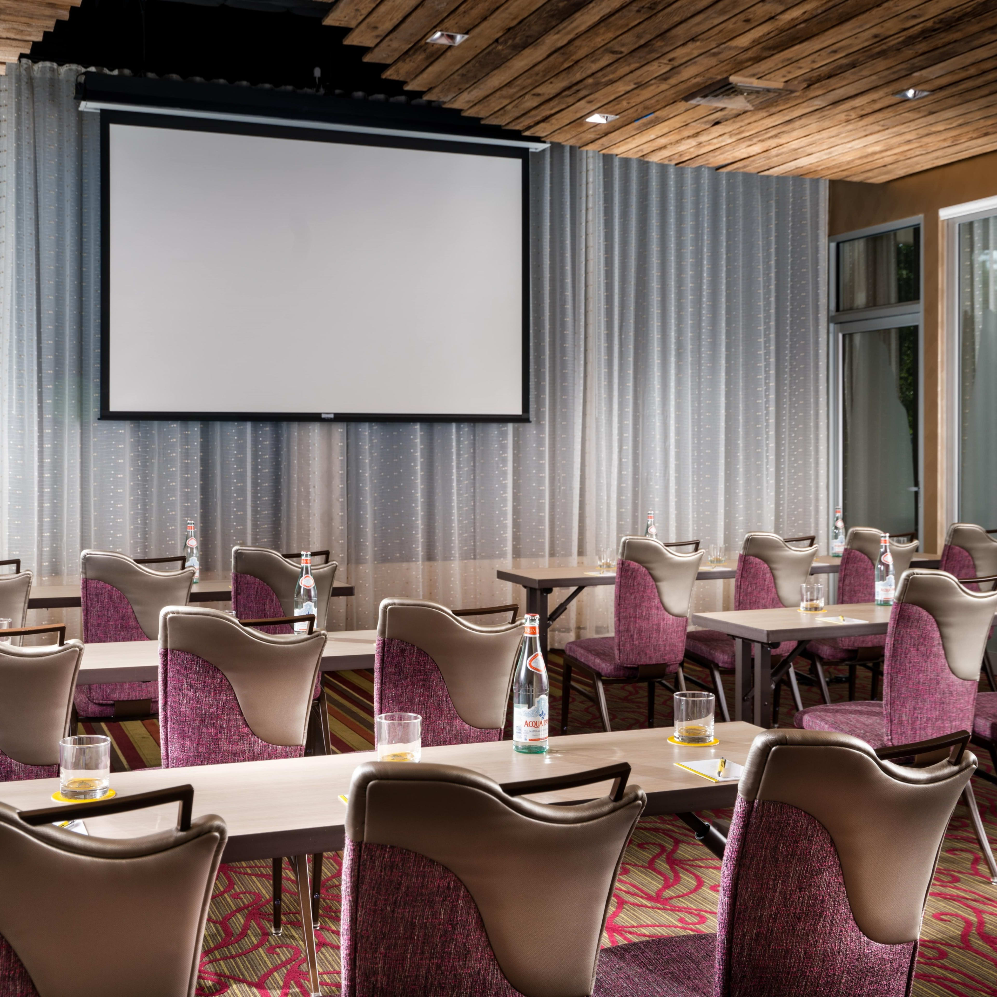 A conference room is set up with rows of tables and chairs facing a large projection screen, with bottled water and glasses on each table.