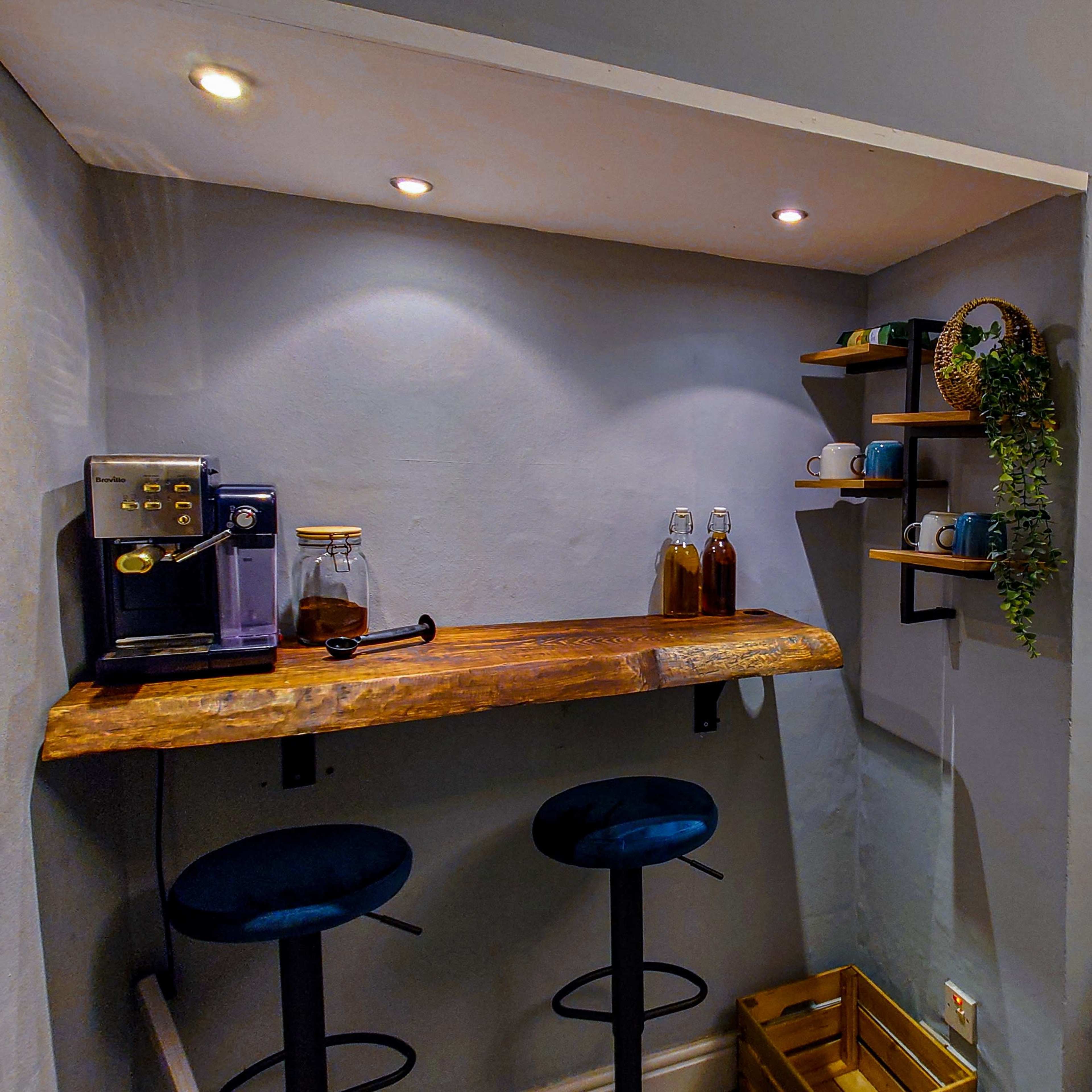 A minimalist corner coffee station featuring a wooden countertop with two bar stools, a coffee machine, glass bottles, cups on a wall shelf, and subtle lighting.