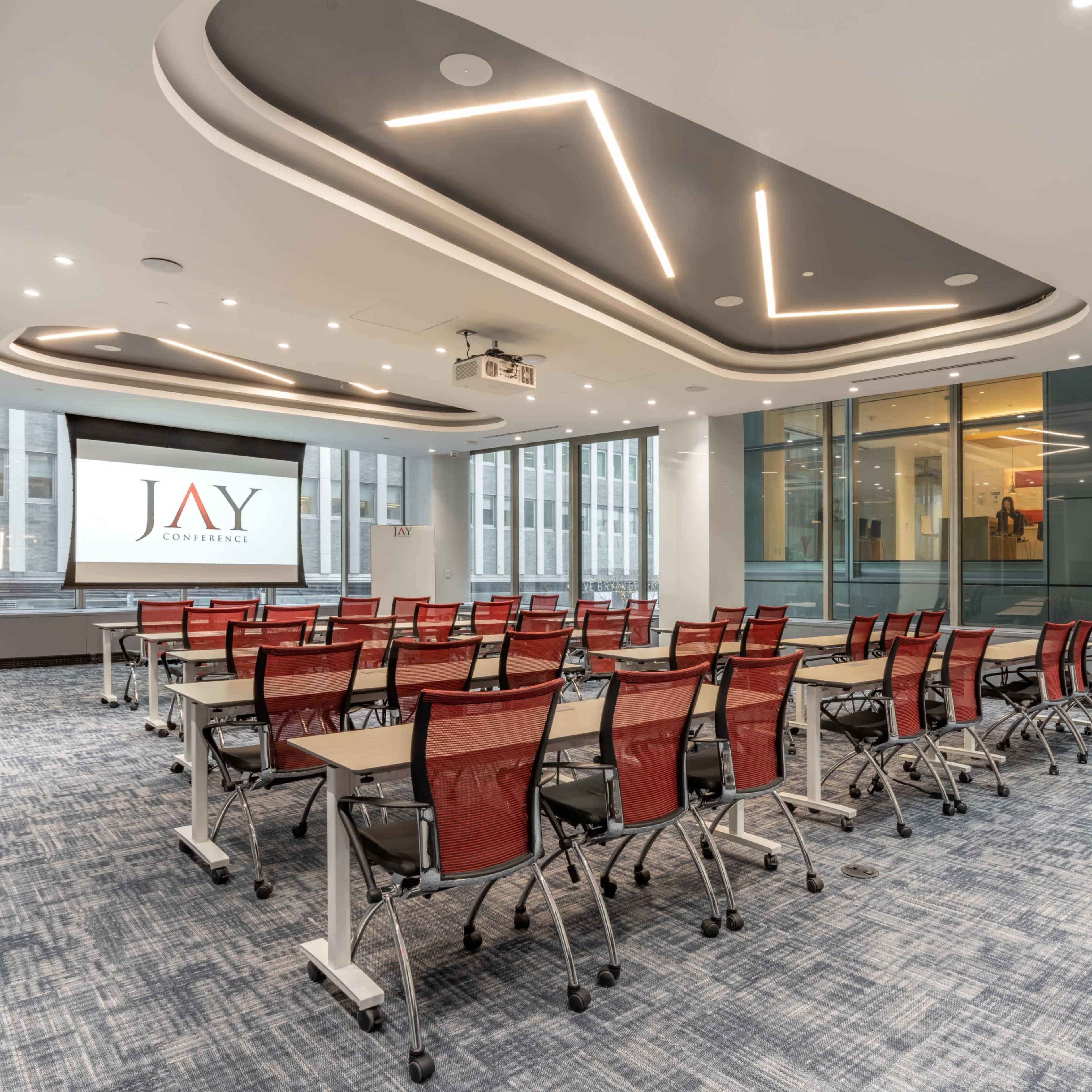 A modern conference room set up with rows of red mesh chairs facing a large screen displaying the logo "JAY" and a spacious layout with windows.