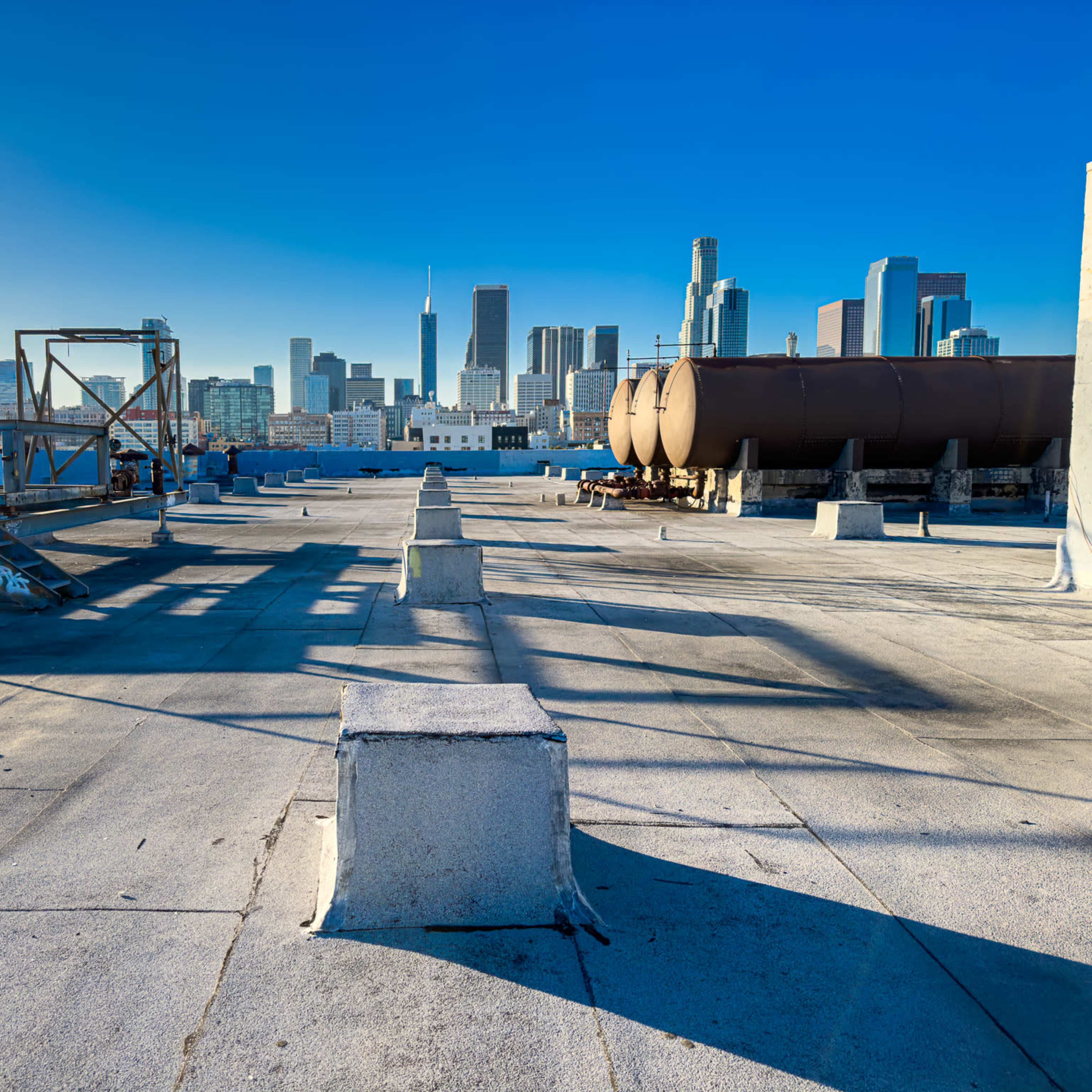 A rooftop area in an urban setting features several concrete blocks and large cylindrical tanks with a skyline of tall buildings in the background.