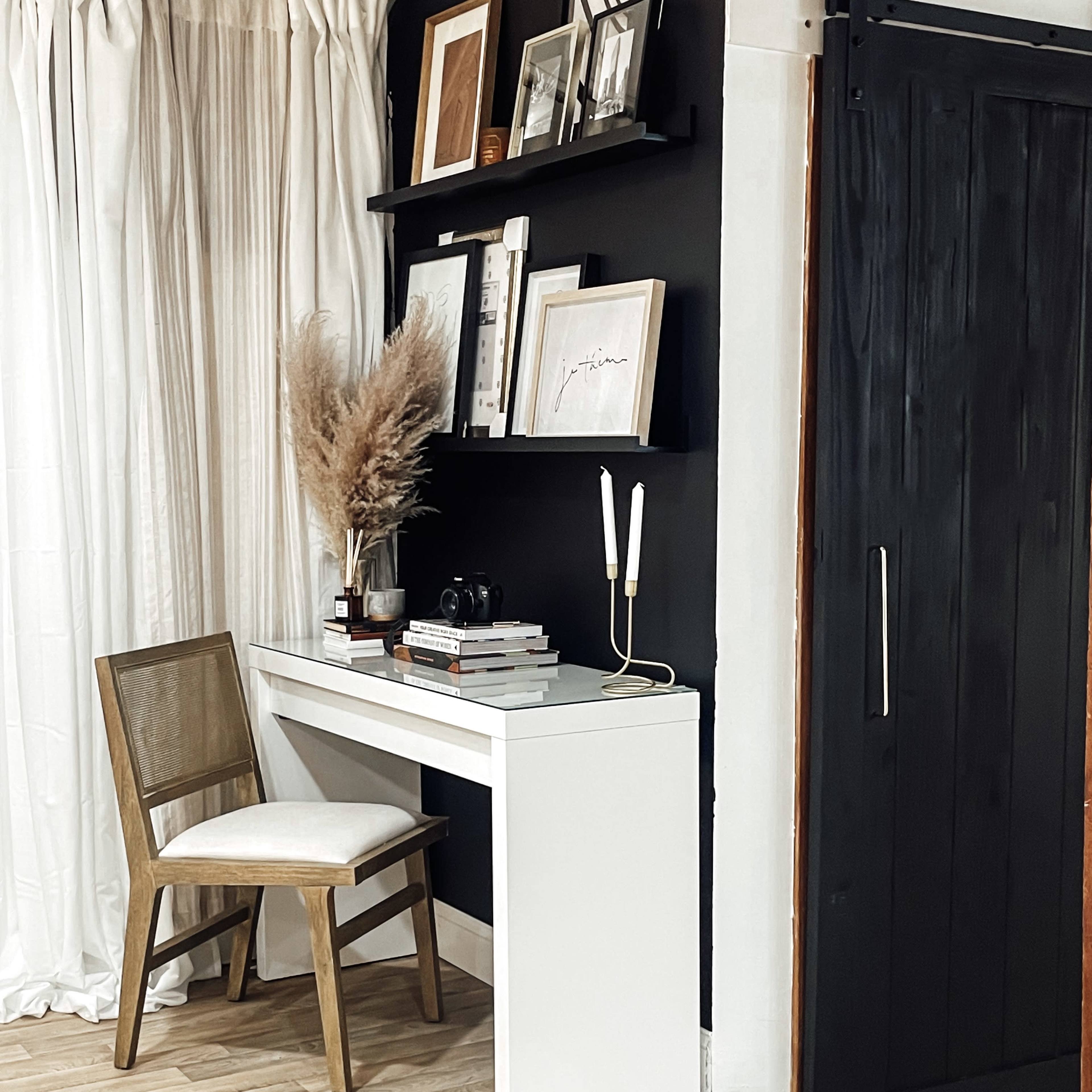 A minimalist workspace features a white desk with a chair, shelves holding framed art, and decor against a dark wall.