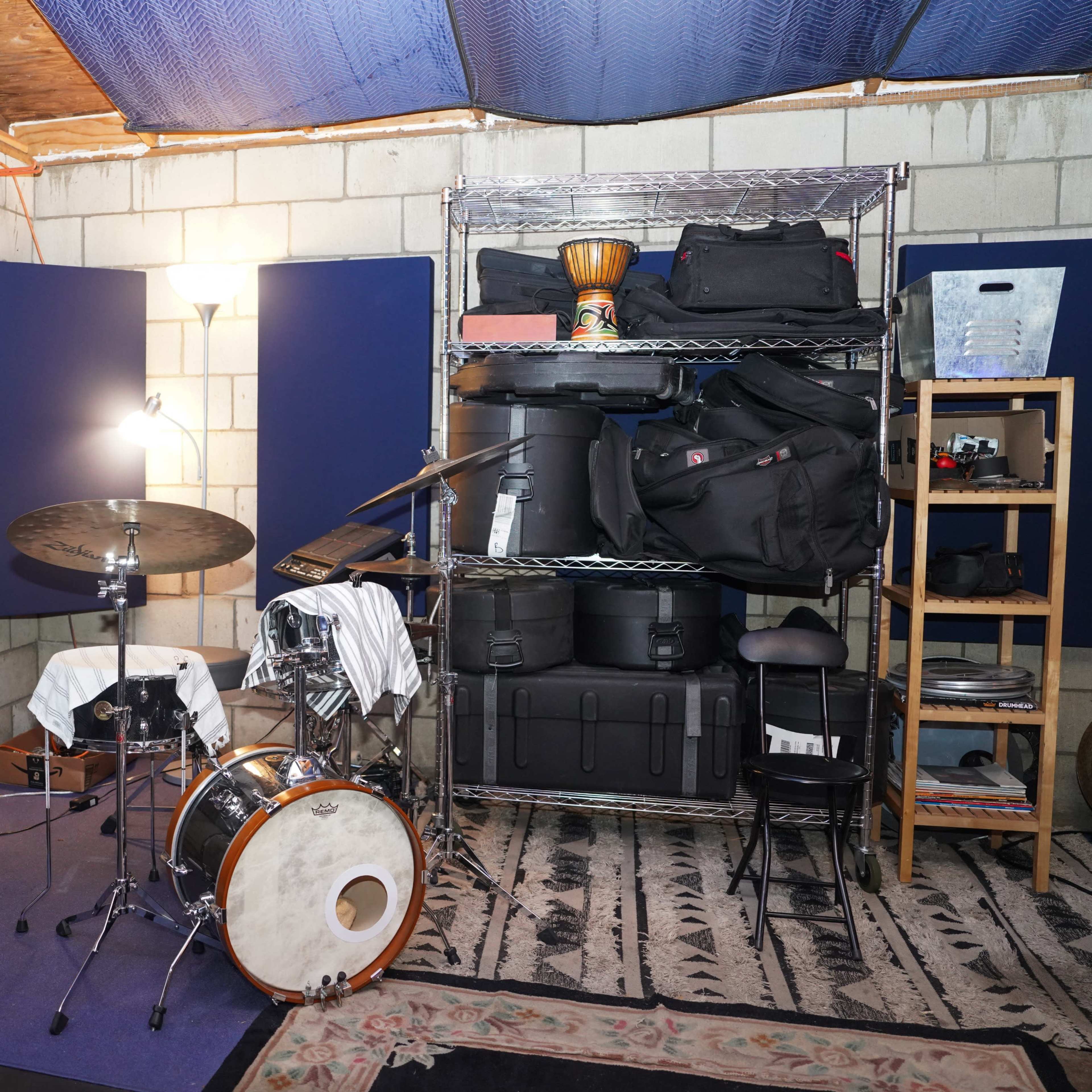 A small music practice room featuring a drum set, amplifiers, and various storage containers organized on shelves.