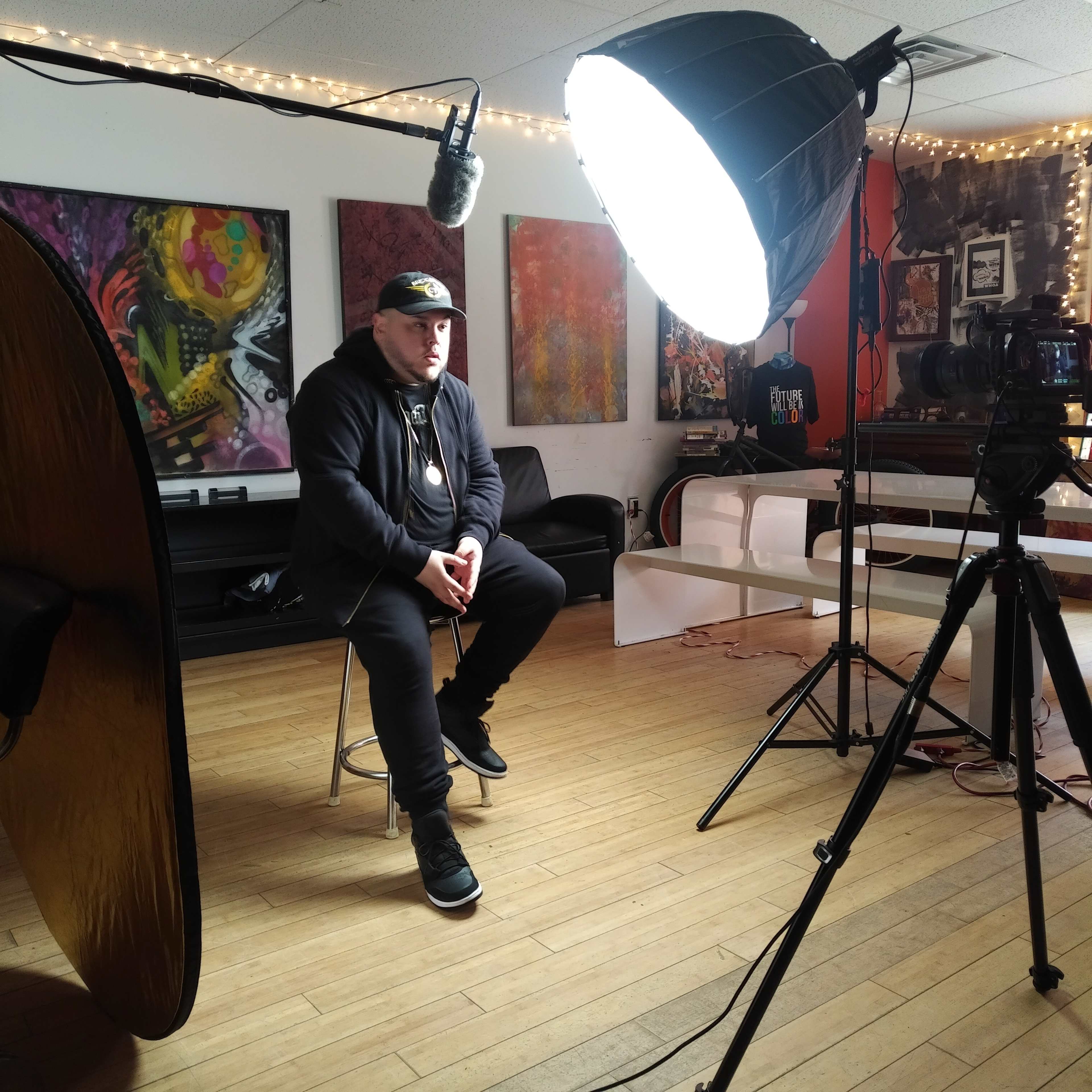 A person sits on a stool in a studio setting, with soft lighting equipment and a camera positioned nearby.